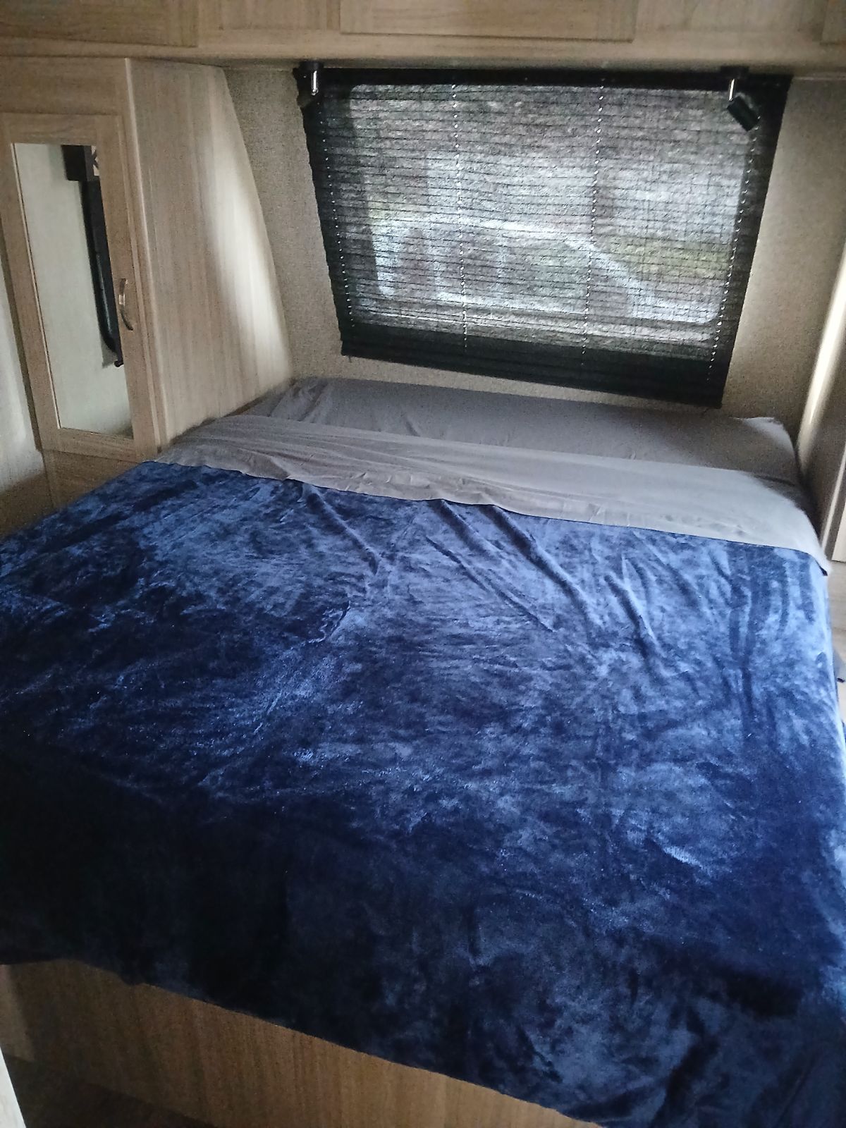 Inside an RV rental: a comfy bed with a plush blue blanket, grey sheets, and window blinds.. Coachmen Other 2021