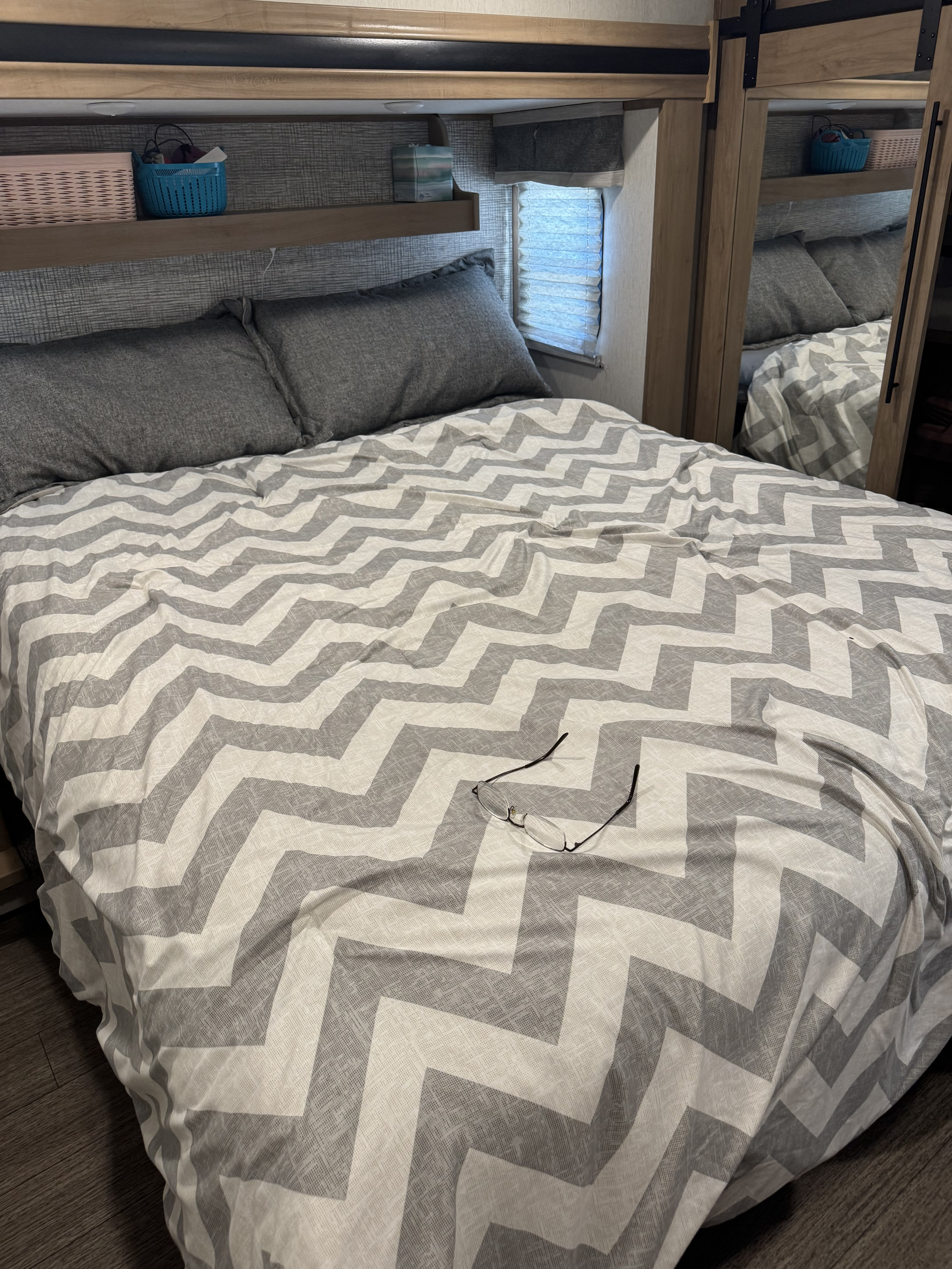 RV rental bedroom: grey pillows, chevron comforter, glasses on bed. Overhead shelf with baskets, window blinds.. Forest River Sabre 2023