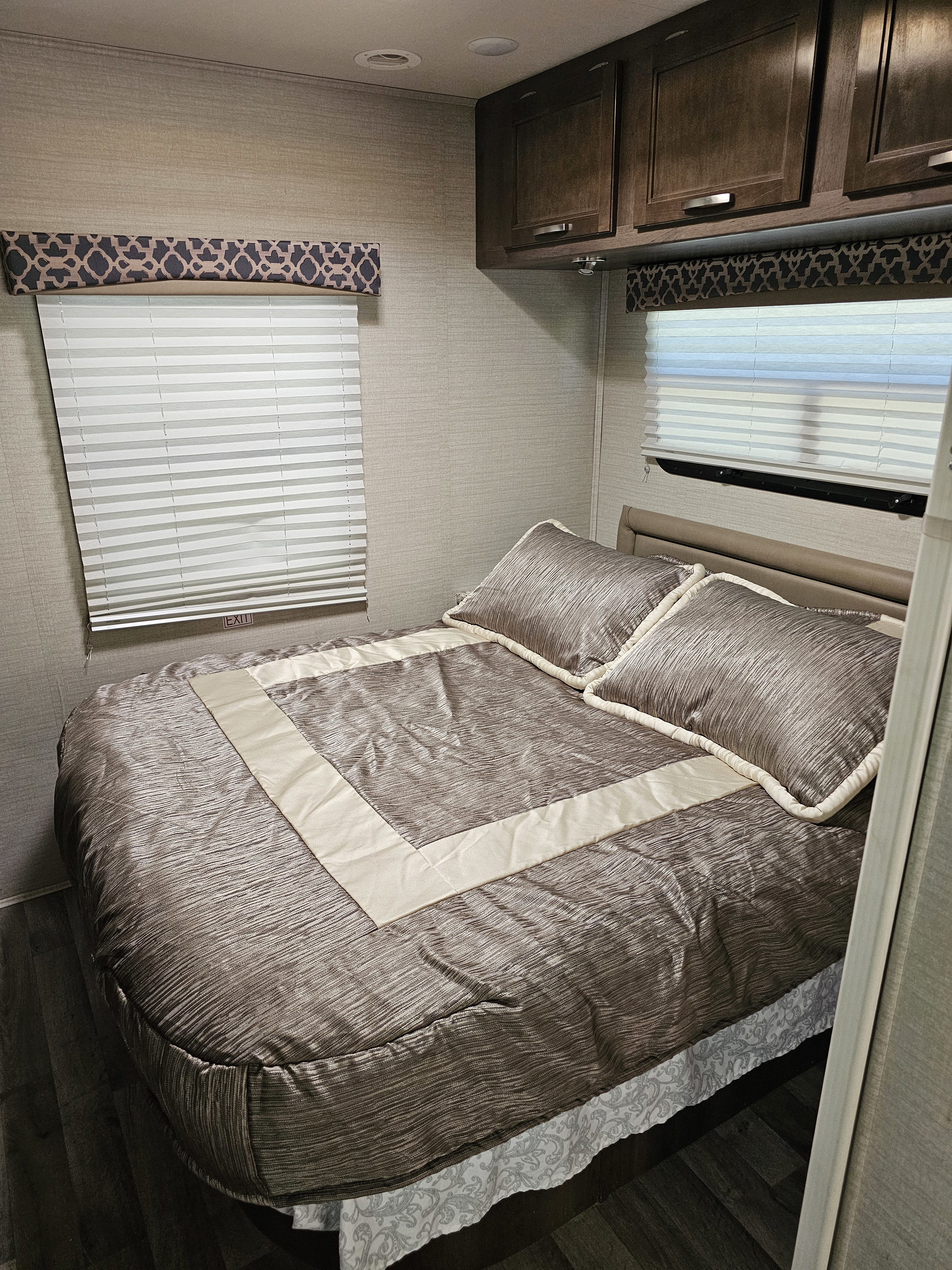 Warm-toned RV rental bedroom with textured brown bedding, sleek overhead cabinets, and windows with light blinds and patterned valances.. Jayco Redhawk 2019