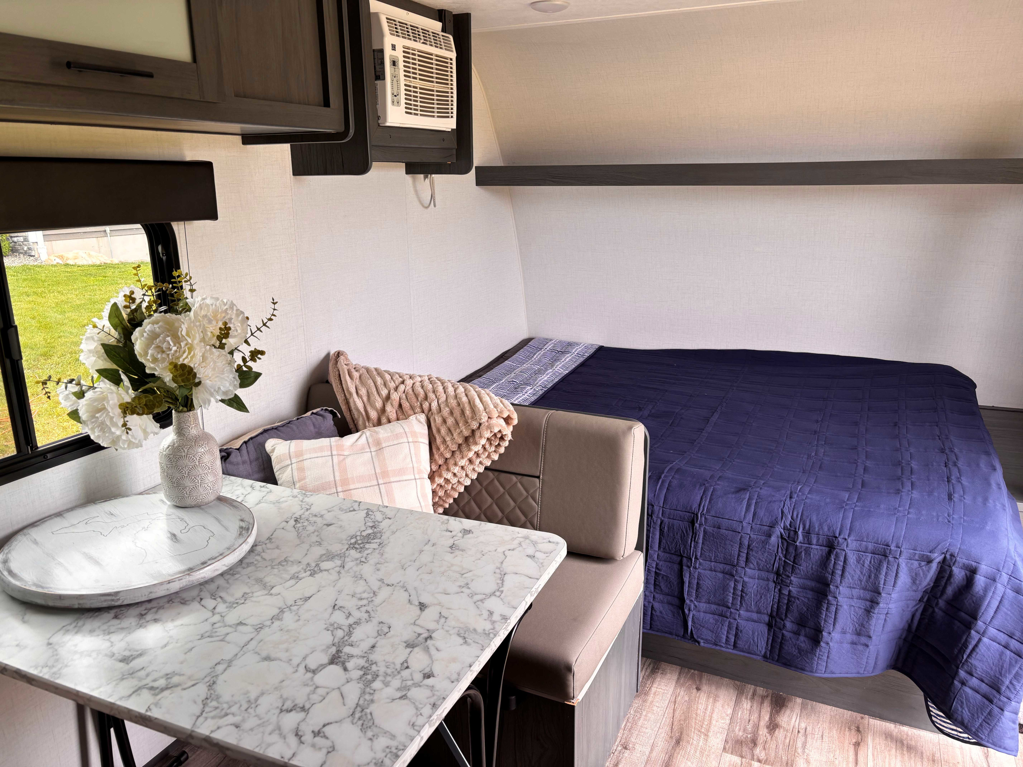 Spacious RV rental interior: a comfy bed with navy quilt, dining area with marble-look table, and cozy seating. Ready for your travels!. Dutchmen Prairie View 2023