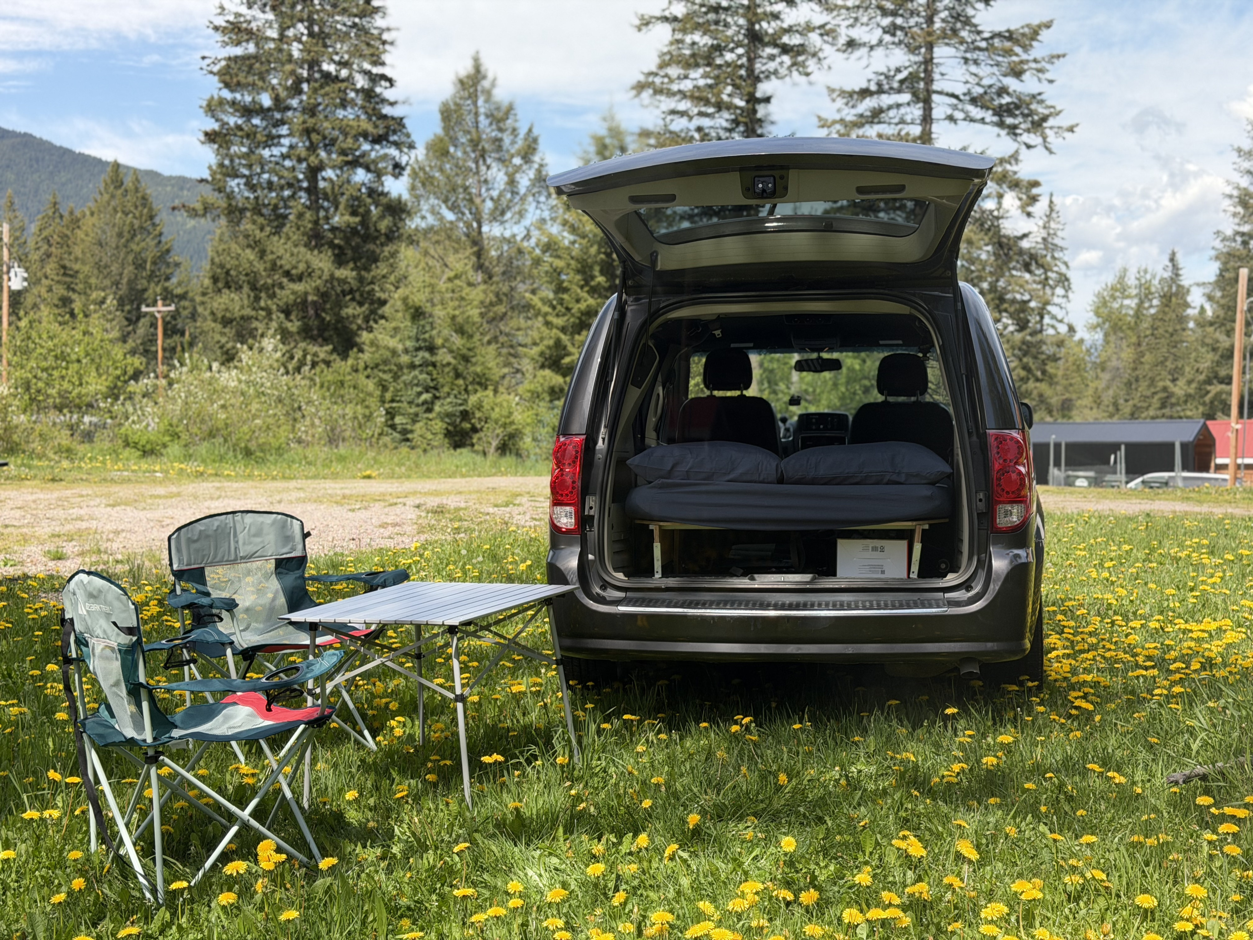 This RV rental offers a perfect van life camping setup. A minivan with a bed, chairs, and table is nestled in a field of dandelions.. Dodge Grand Caravan 2017