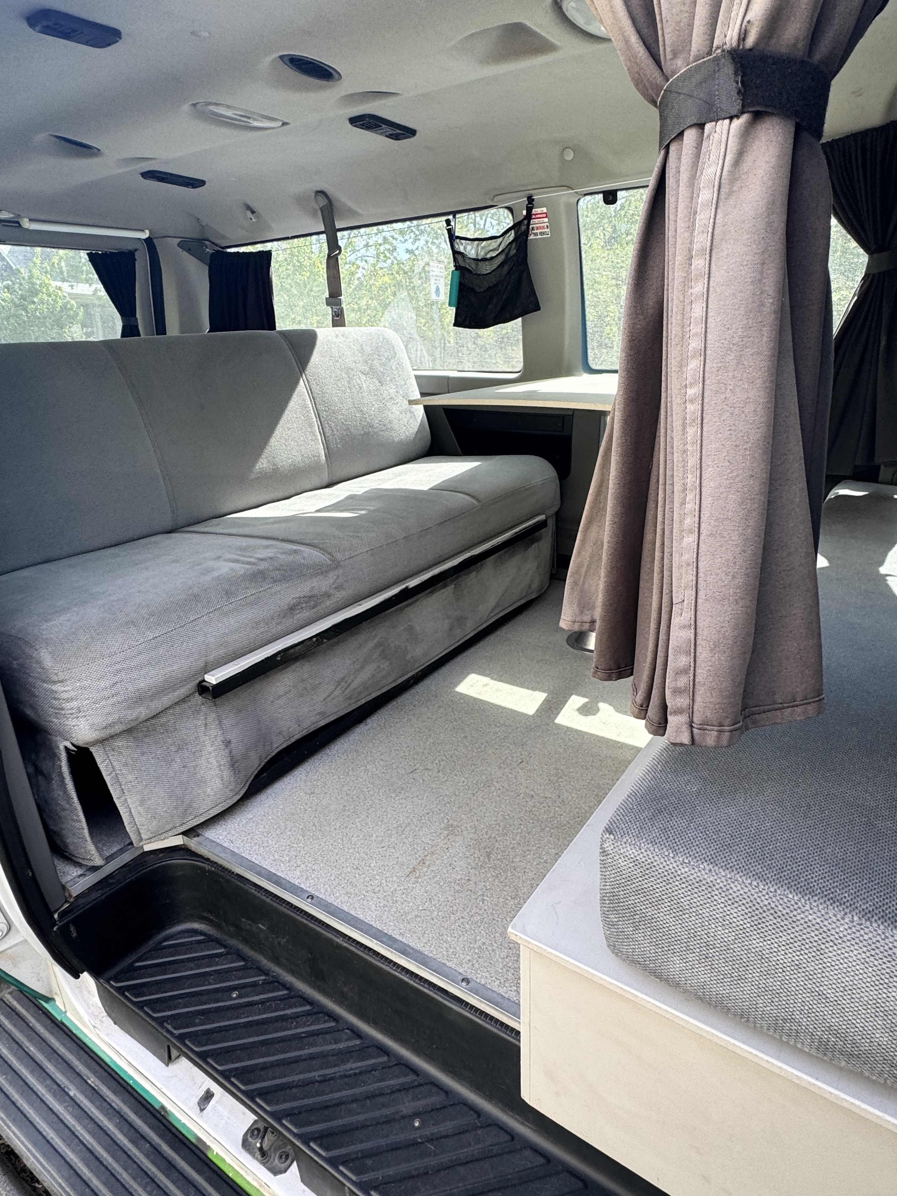 Cozy RV rental interior: grey bench seat (converts to bed), small table, & privacy curtains, ready for your road trip.. Ford Econoline 150 2012