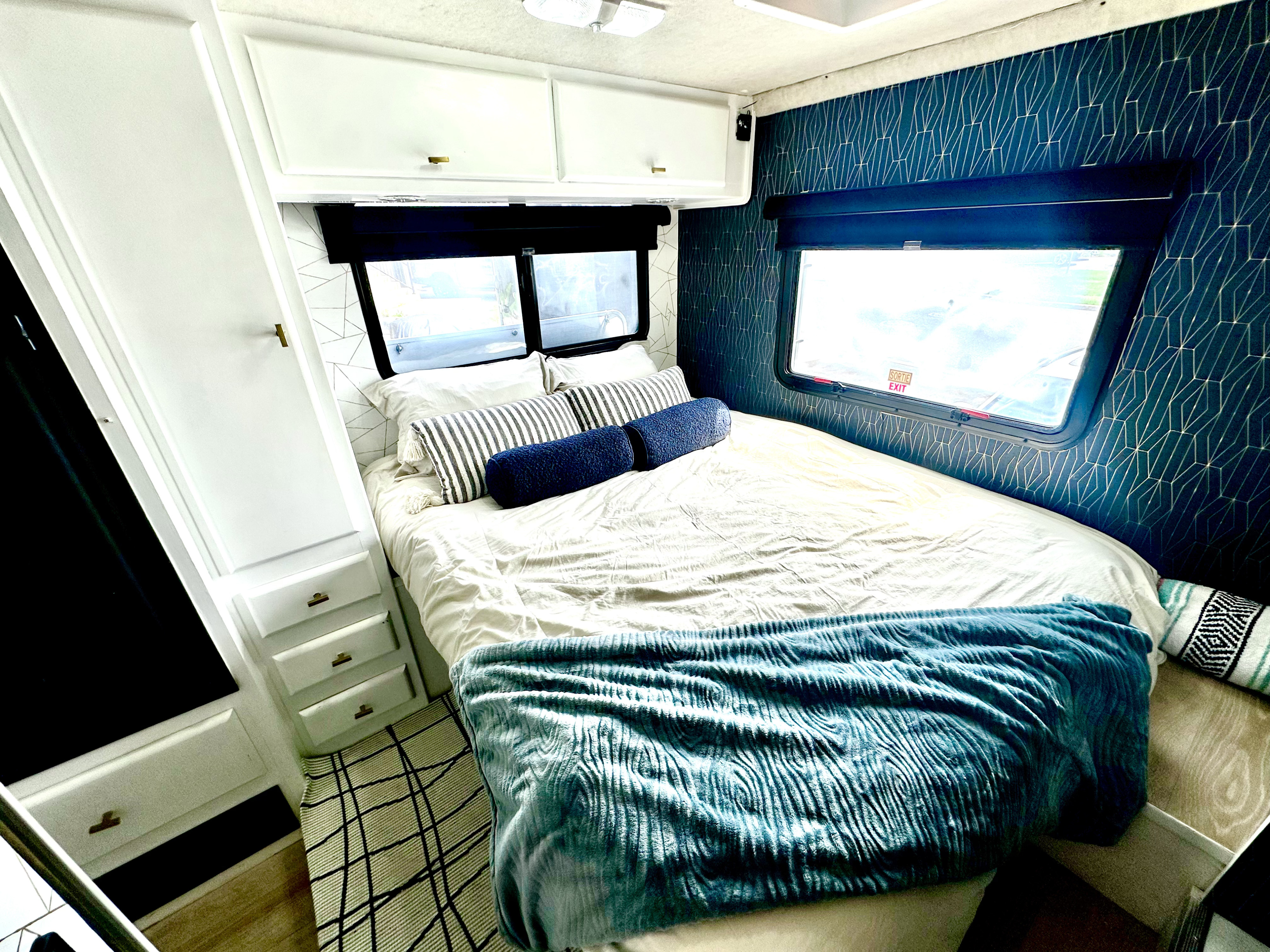 Inside a cozy RV rental bedroom: a comfortable bed with a blue blanket, white built-in cabinets, and stylish geometric wallpaper.. Jamboree Sport 2009