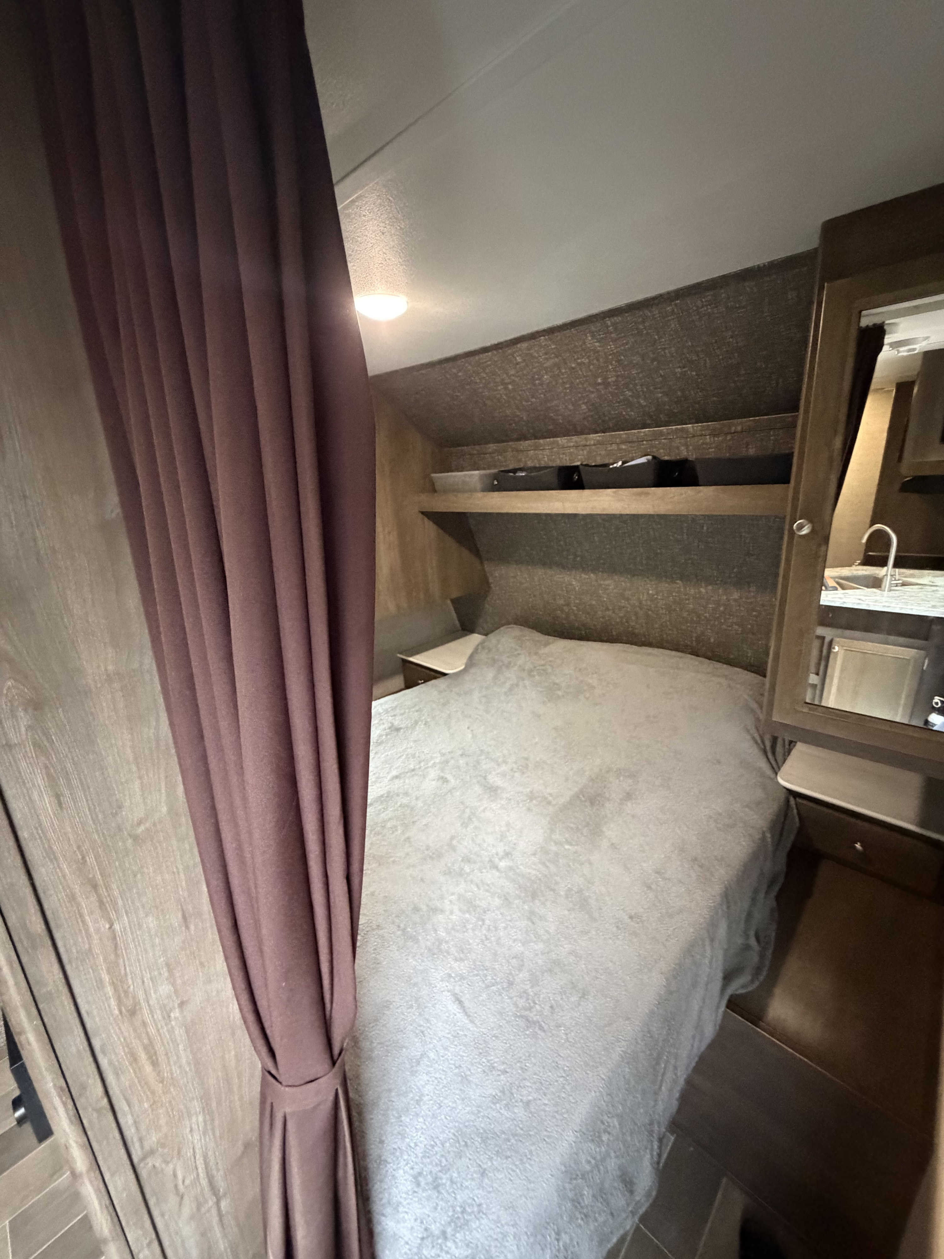 Inside a cozy RV rental: a bed with shelves, a brown curtain, and a mirrored reflection of a small kitchen.. Coleman 295QB 2019