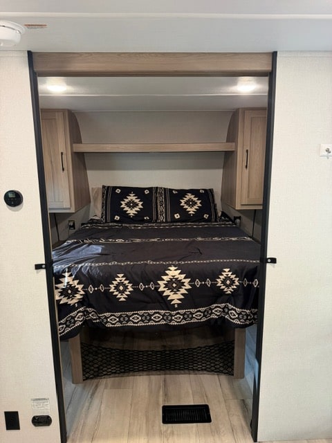 RV rental bedroom interior, showing a comfortable bed with patterned bedding, overhead storage, and light wood flooring.. Keystone RV Coleman Light 2025