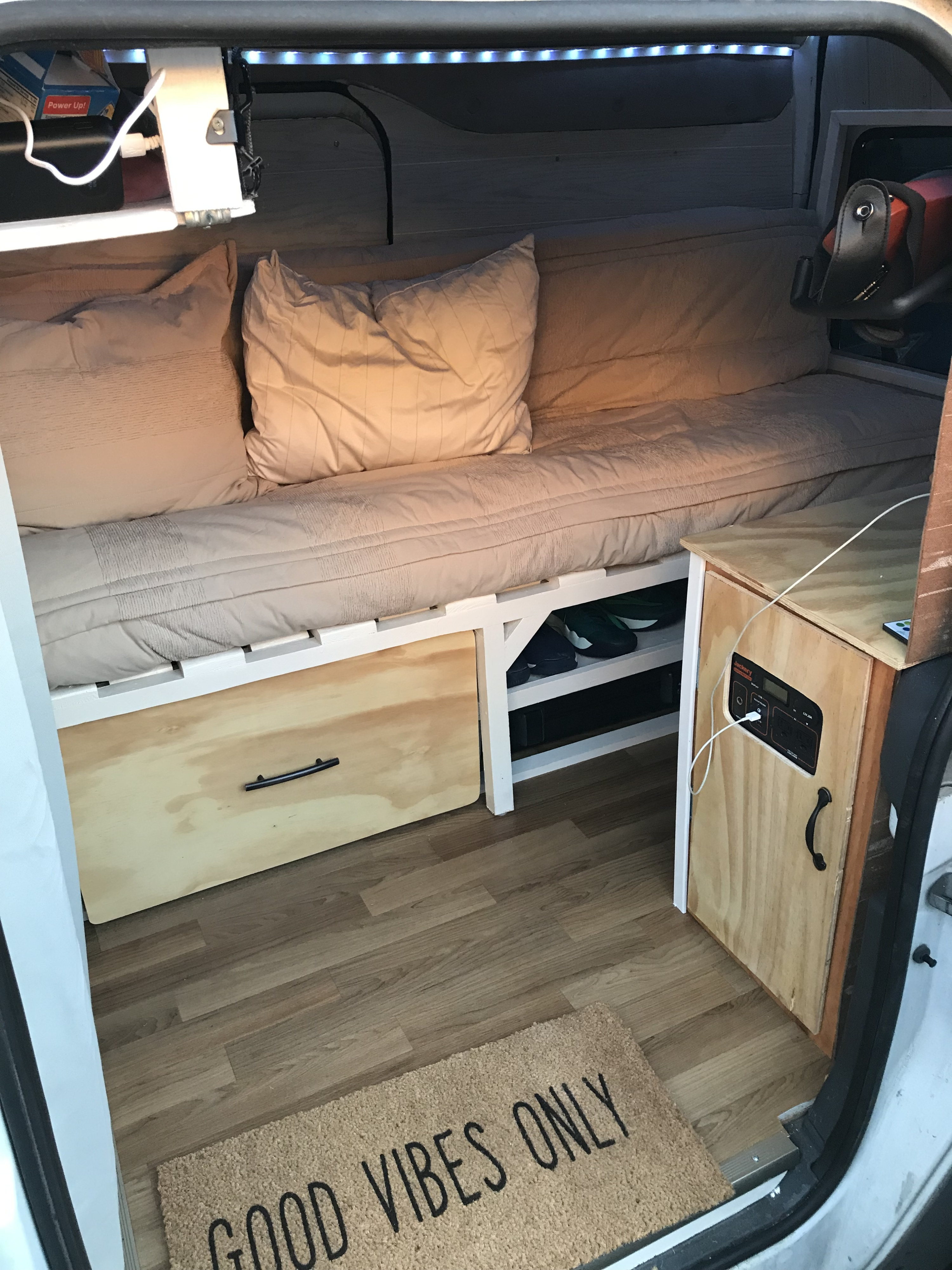 Cozy RV rental interior with a bed, under-bed storage, power station, and a "GOOD VIBES ONLY" welcome mat.. Ford Transit 2011