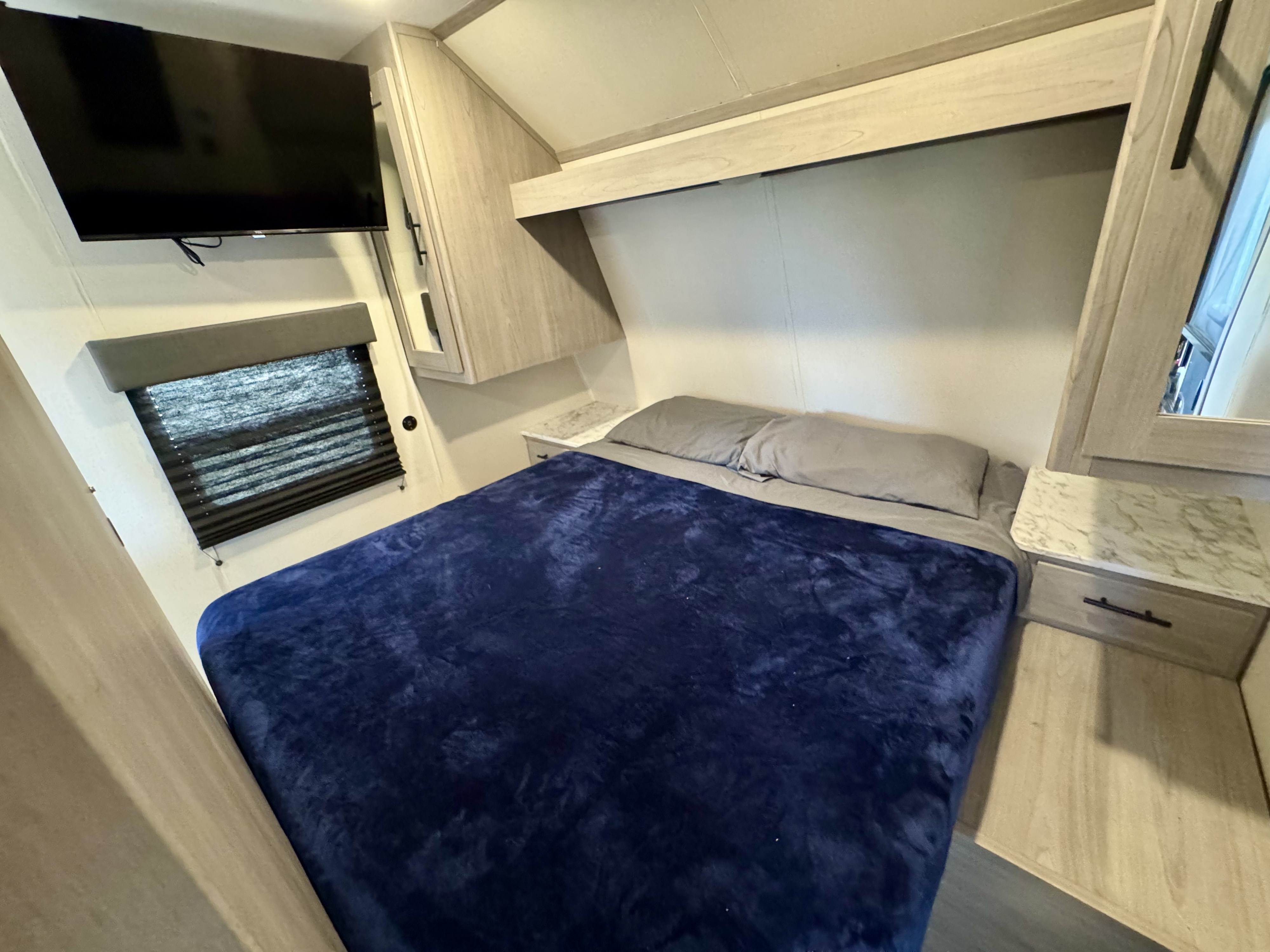 Cozy RV rental bedroom featuring a bed with a blue blanket, mounted TV, window, and built-in storage.. Forest River Cherokee Grey Wolf 2025