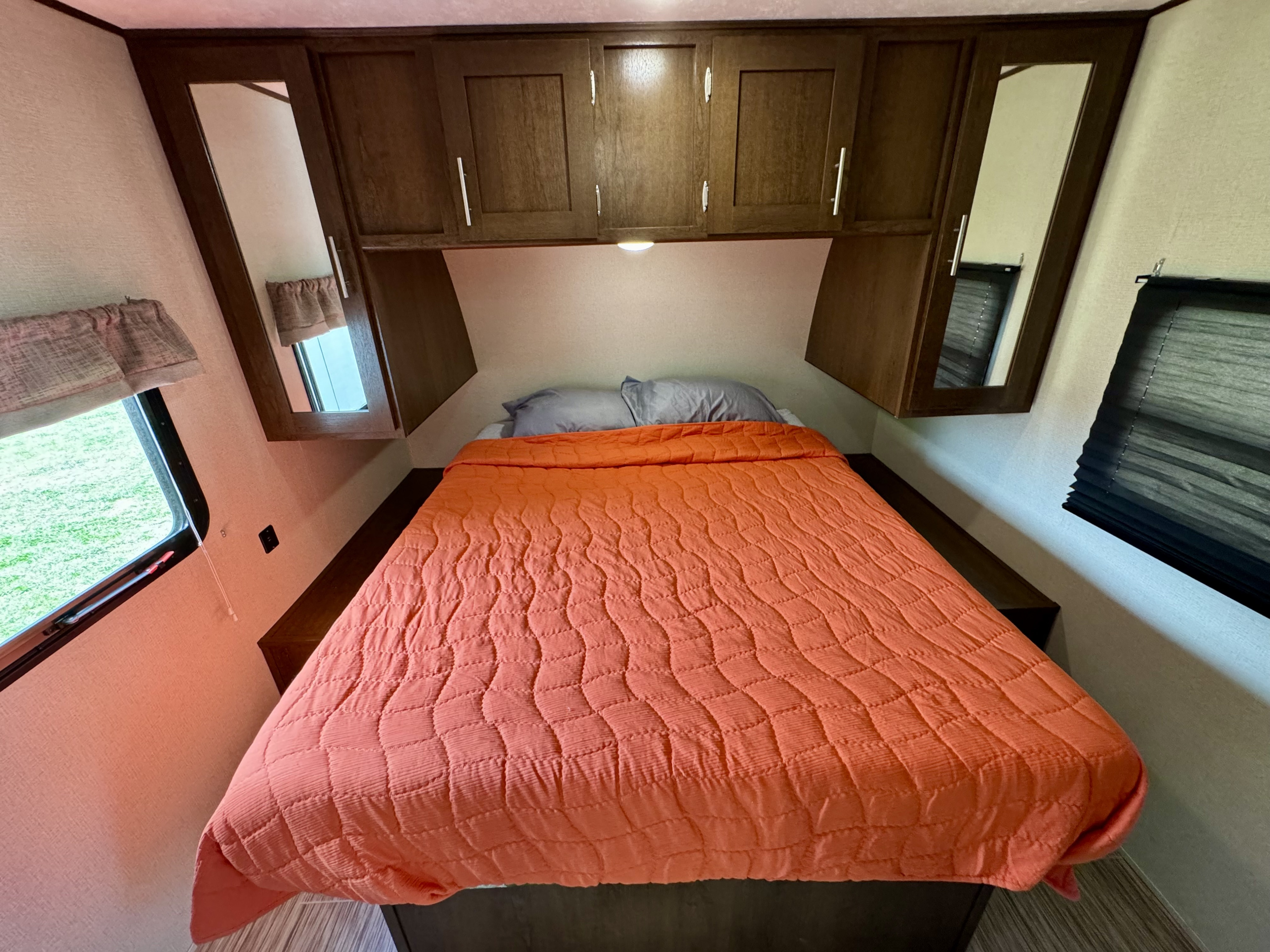 Cozy rv rental bedroom features an orange comforter, grey pillows, overhead cabinets, mirrors, and windows.. Avenger ATI 2020