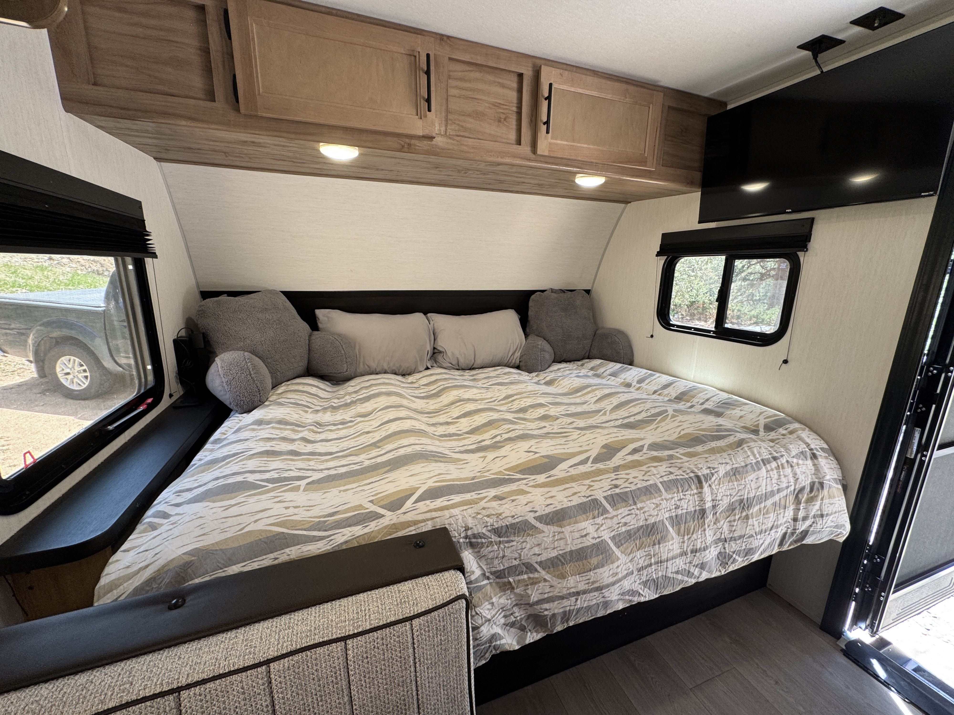 This RV rental bedroom features a cozy bed with patterned bedding, overhead cabinets, a TV, and windows showing the outside.. Forest River Cherokee Wolf Pup 2025