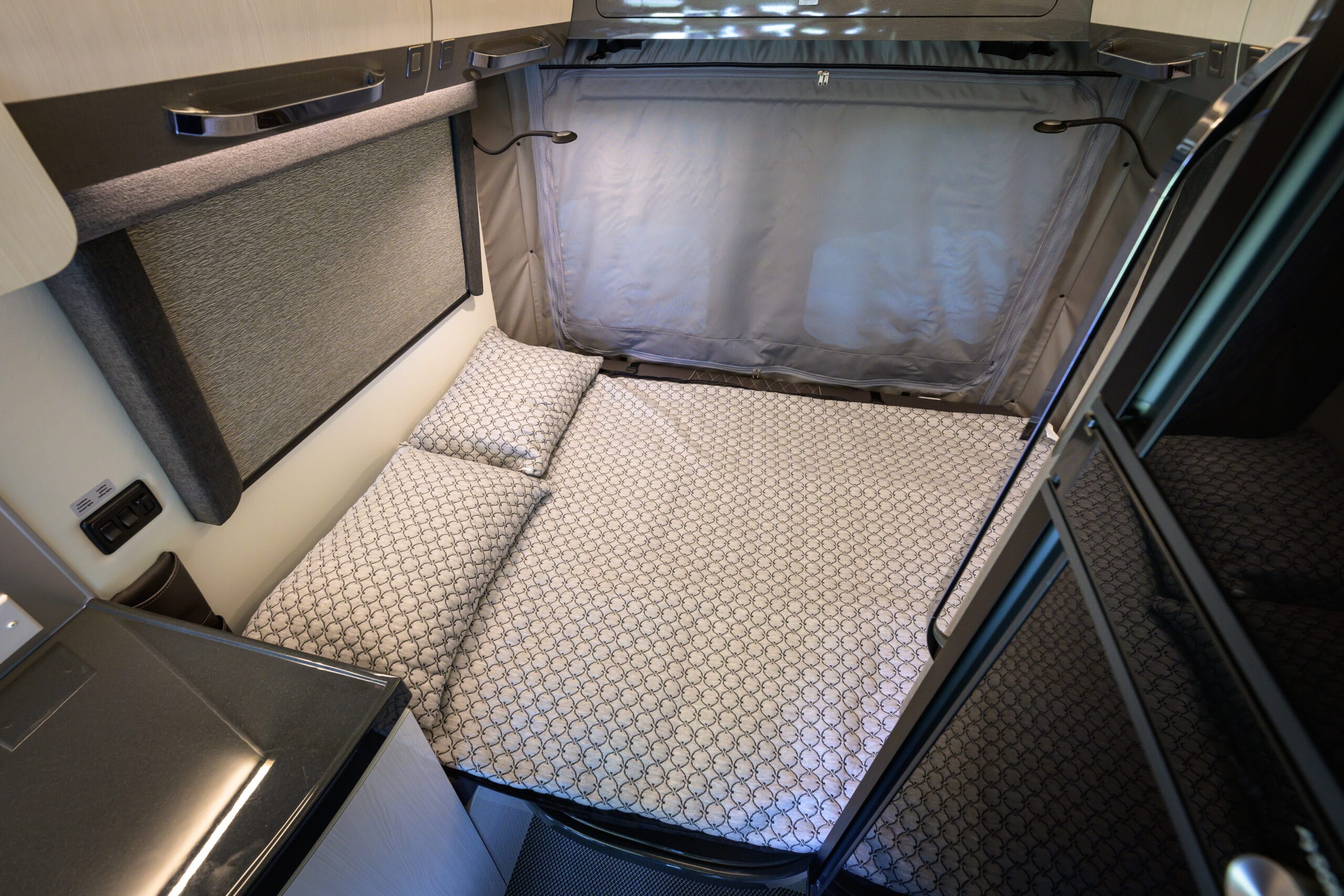 Interior of an rv rental: a compact sleeping area with patterned bed, pillows, overhead storage, and an adjustable reading light.. Other Gala Montecarlo 2025