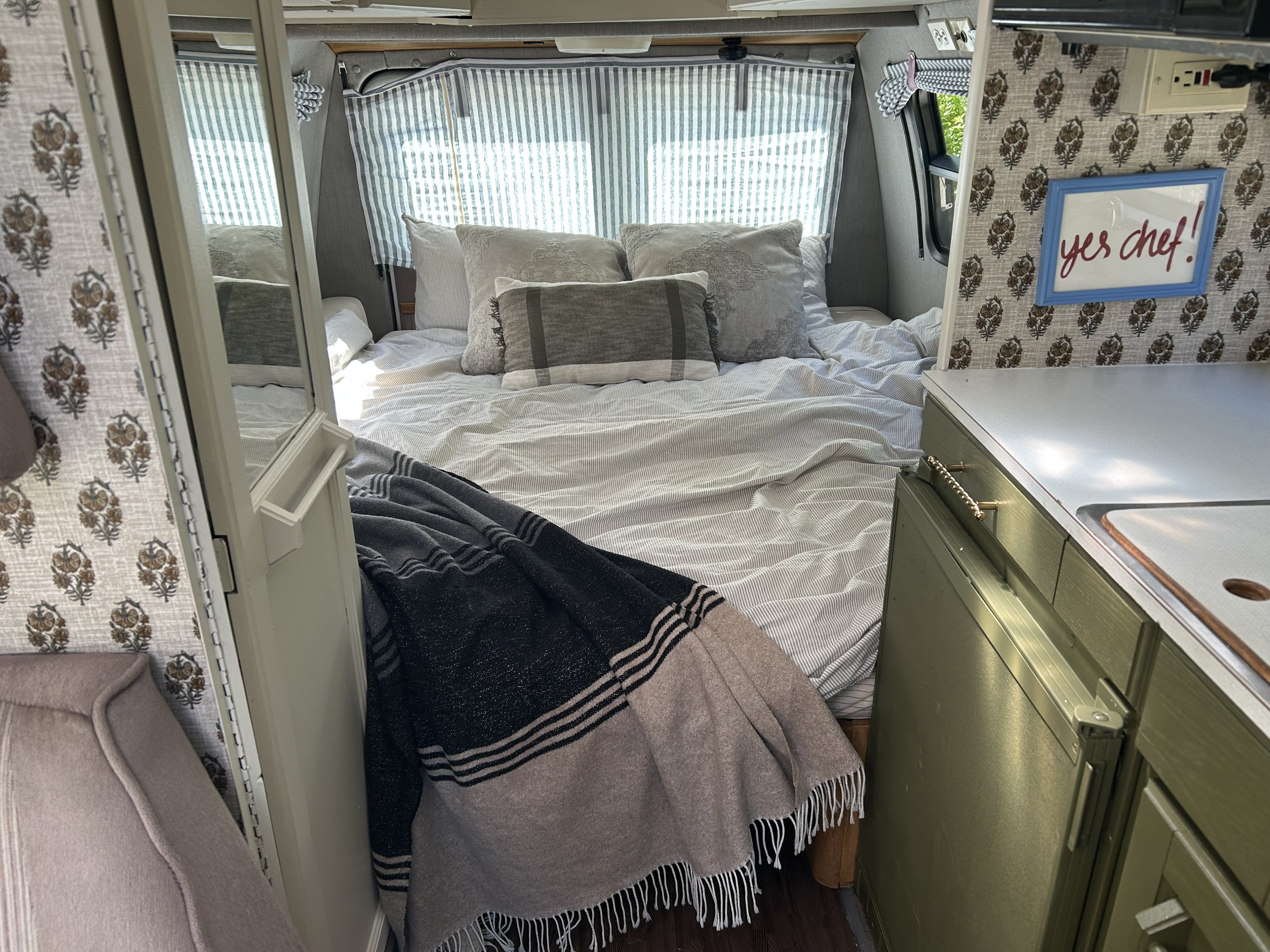 Cozy RV rental interior showcasing a comfortable bed with striped bedding, curtains, and a compact kitchen with a "yes chef!" sign.. Roadtrek 190 Popular 1995
