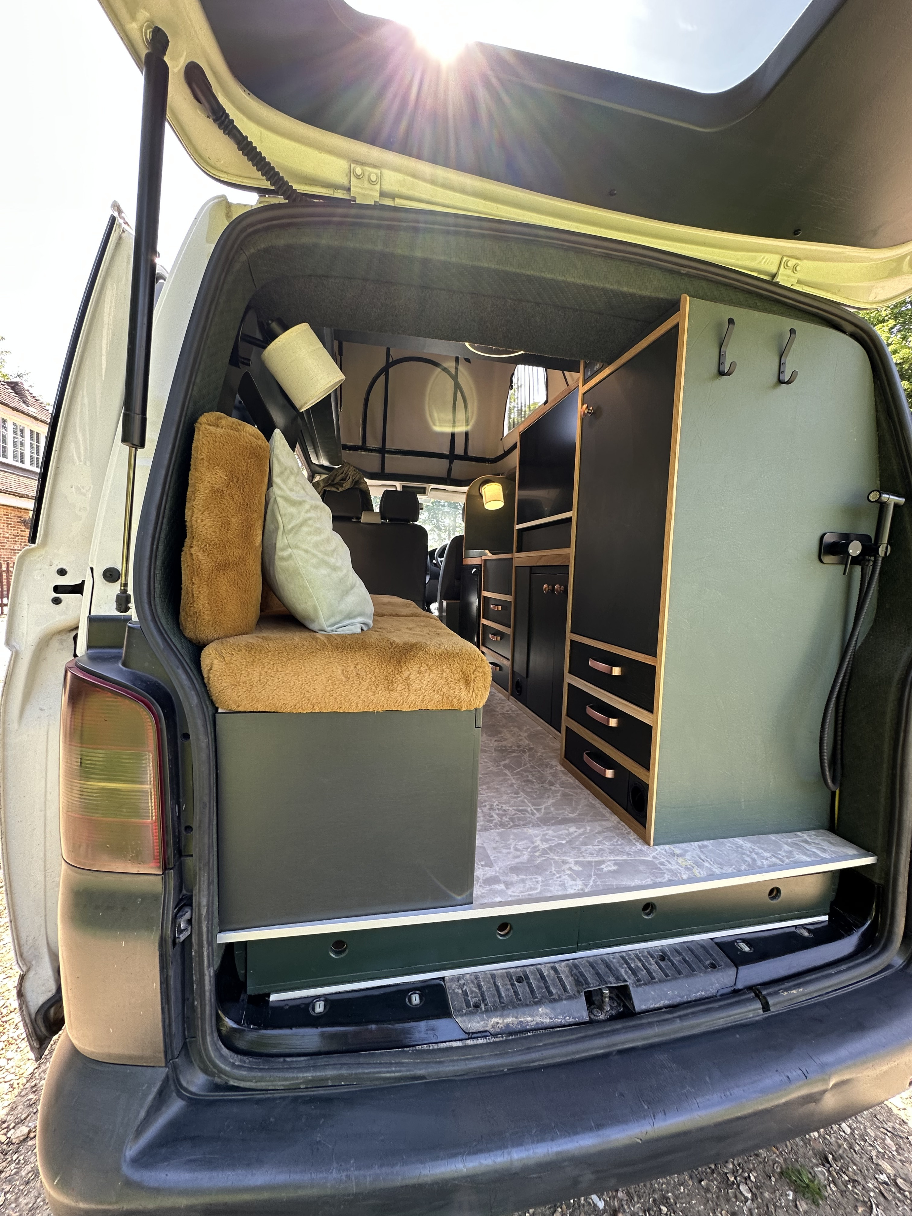 Inside a custom rv rental camper van: plush bed, green & black cabinets, and a pop-top, seen from the open tailgate on a sunny day.. Volkswagen transporter t5.1 2012