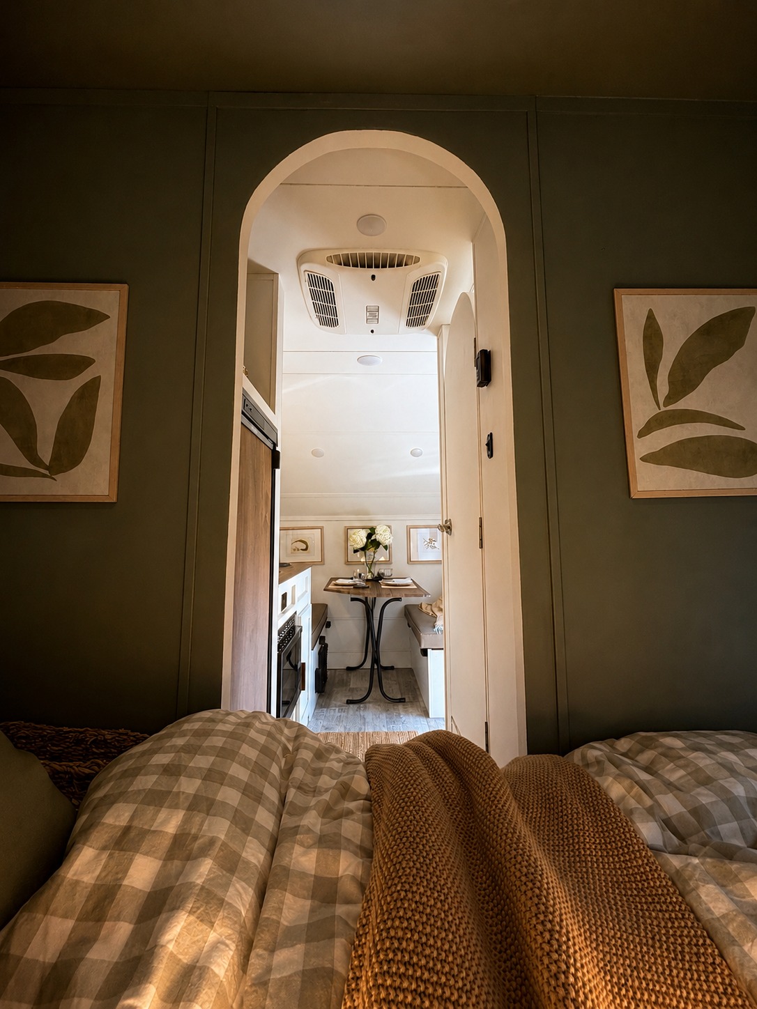 Cozy RV rental: Olive walls, checkered bedding, and archway to a compact dining area with a small table and benches.. R-Pod R-Pod Trailer 2024