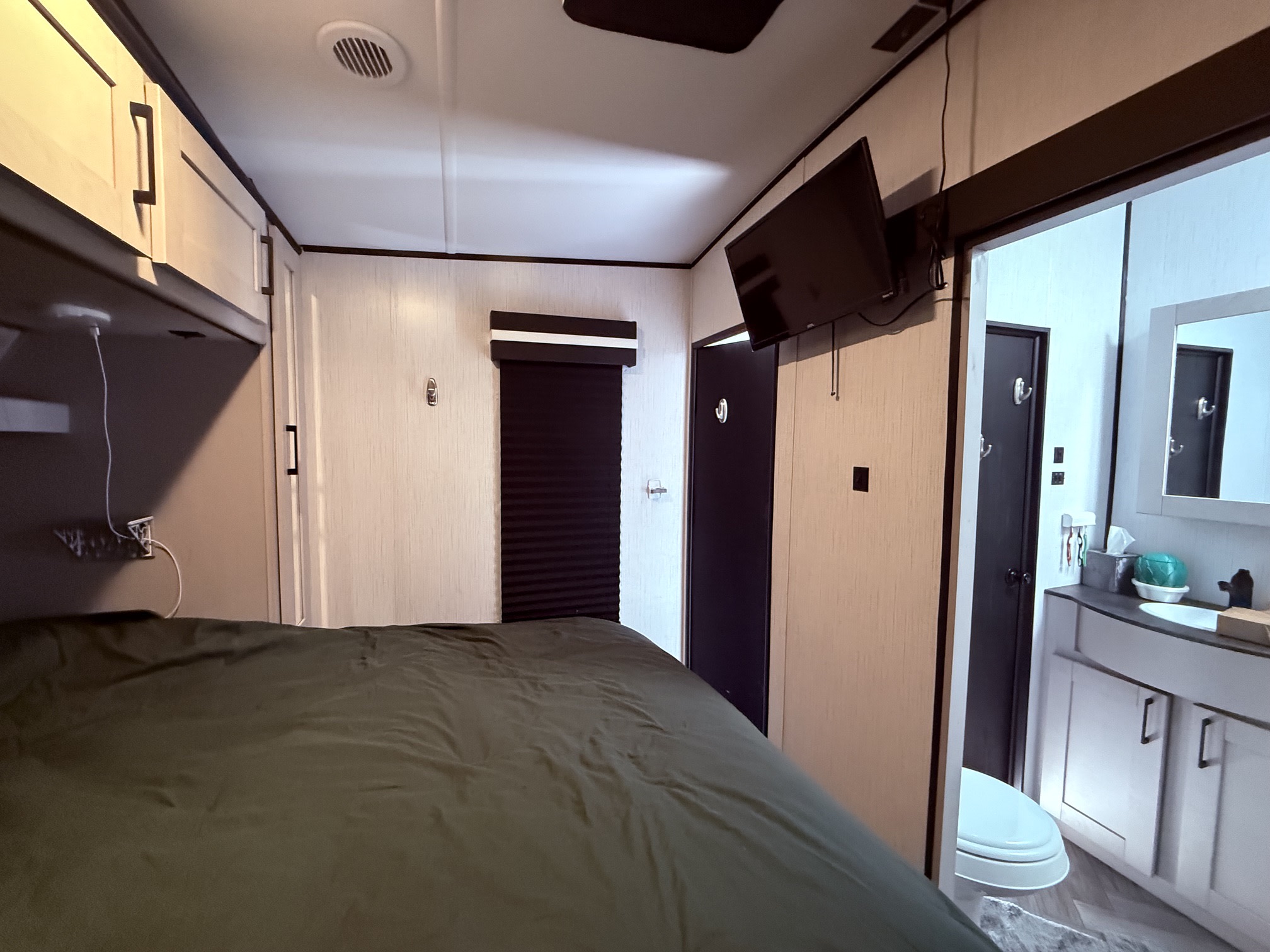 Interior of an RV rental bedroom with a green bed, overhead cabinets, wall-mounted TV, and an open doorway to a bathroom.. Heartland RVs Torque Toy Hauler 2022