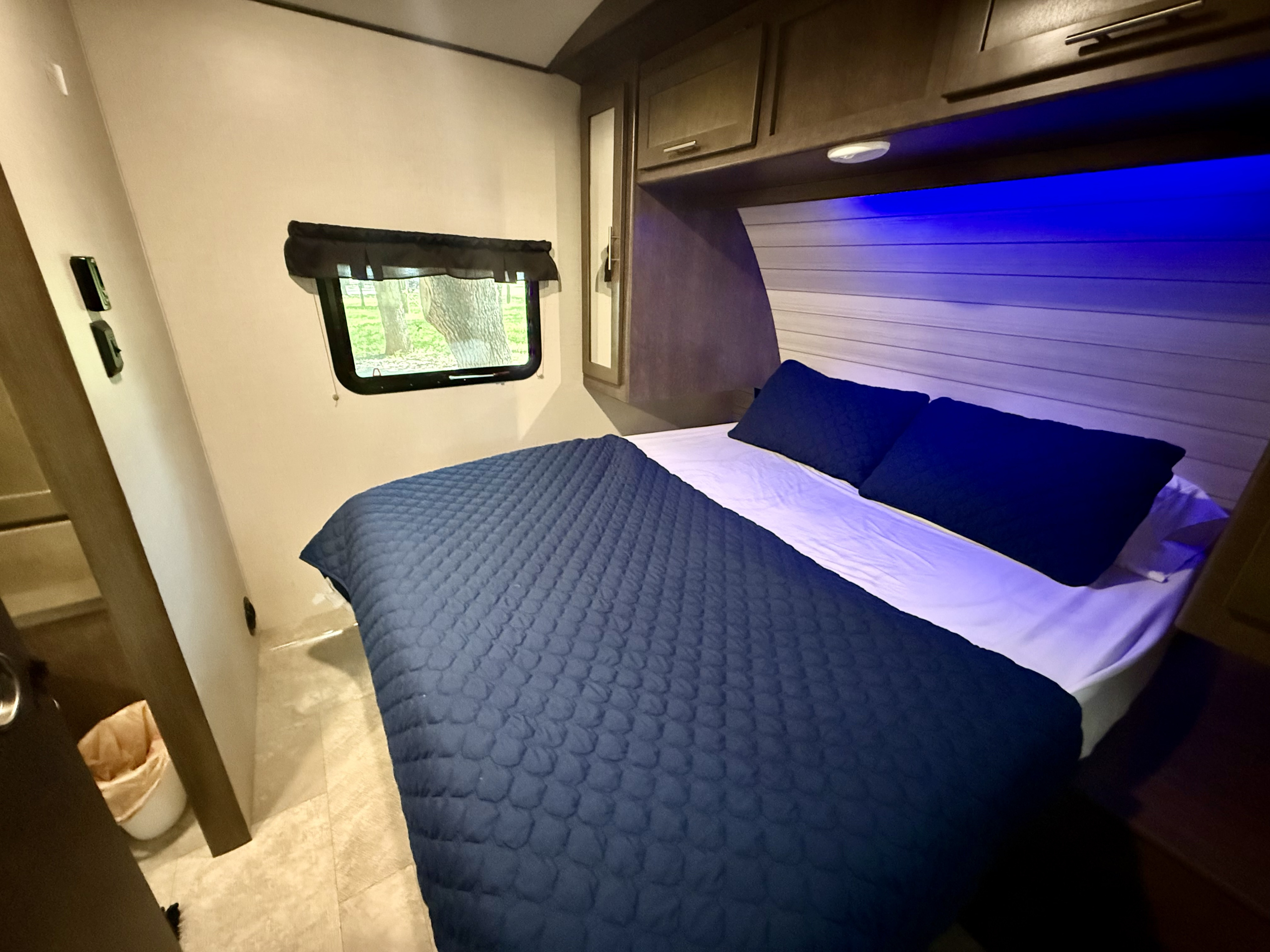 Cozy RV rental bedroom featuring a bed with blue comforter, pillows, white sheets, ambient blue lighting, and a window view.. Crossroads RV Sunset Trail Super Lite 2018