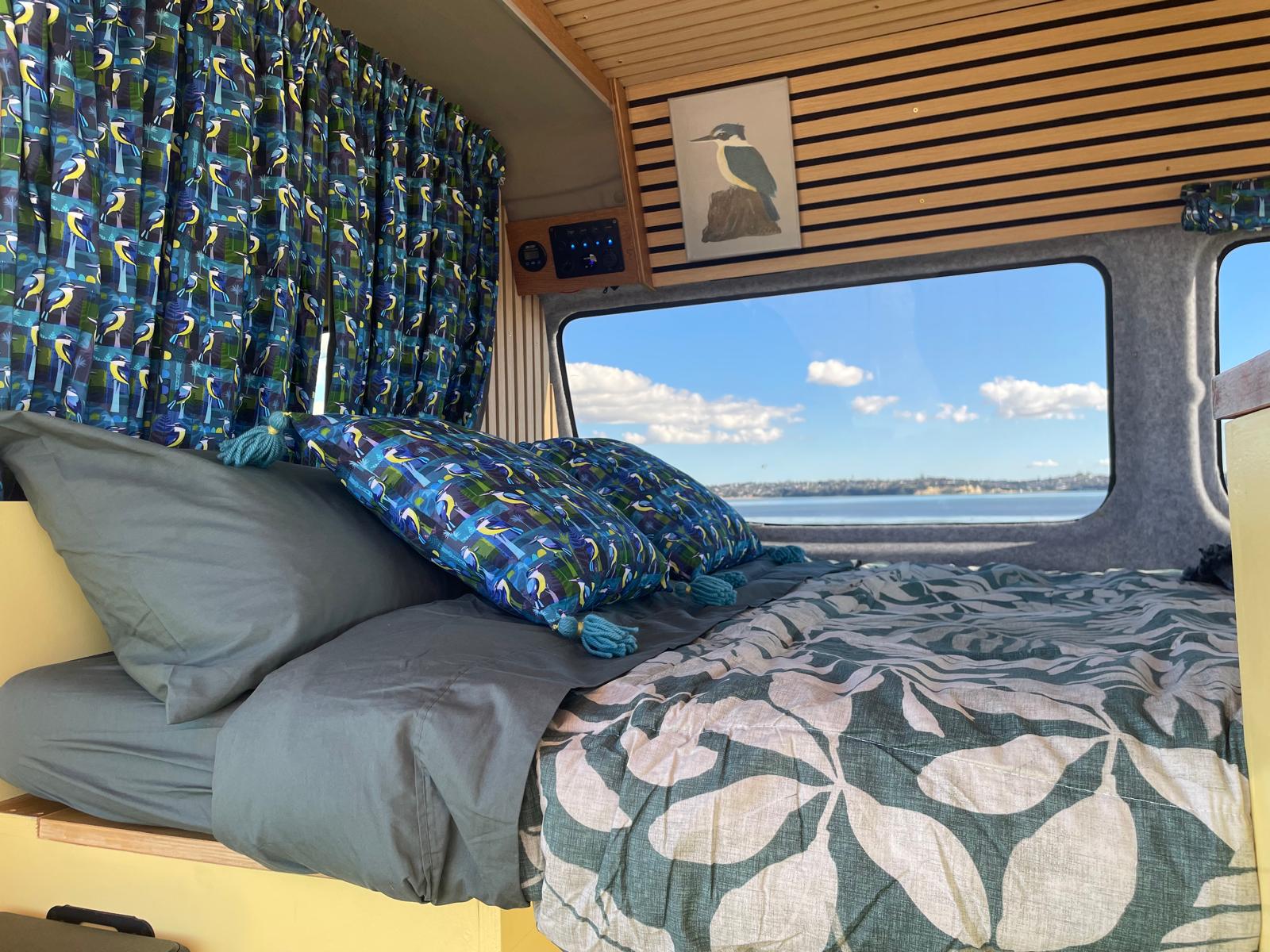 An rv rental's cozy bed with vibrant bird-themed decor and a picturesque ocean view out the window.. Other Other 2019