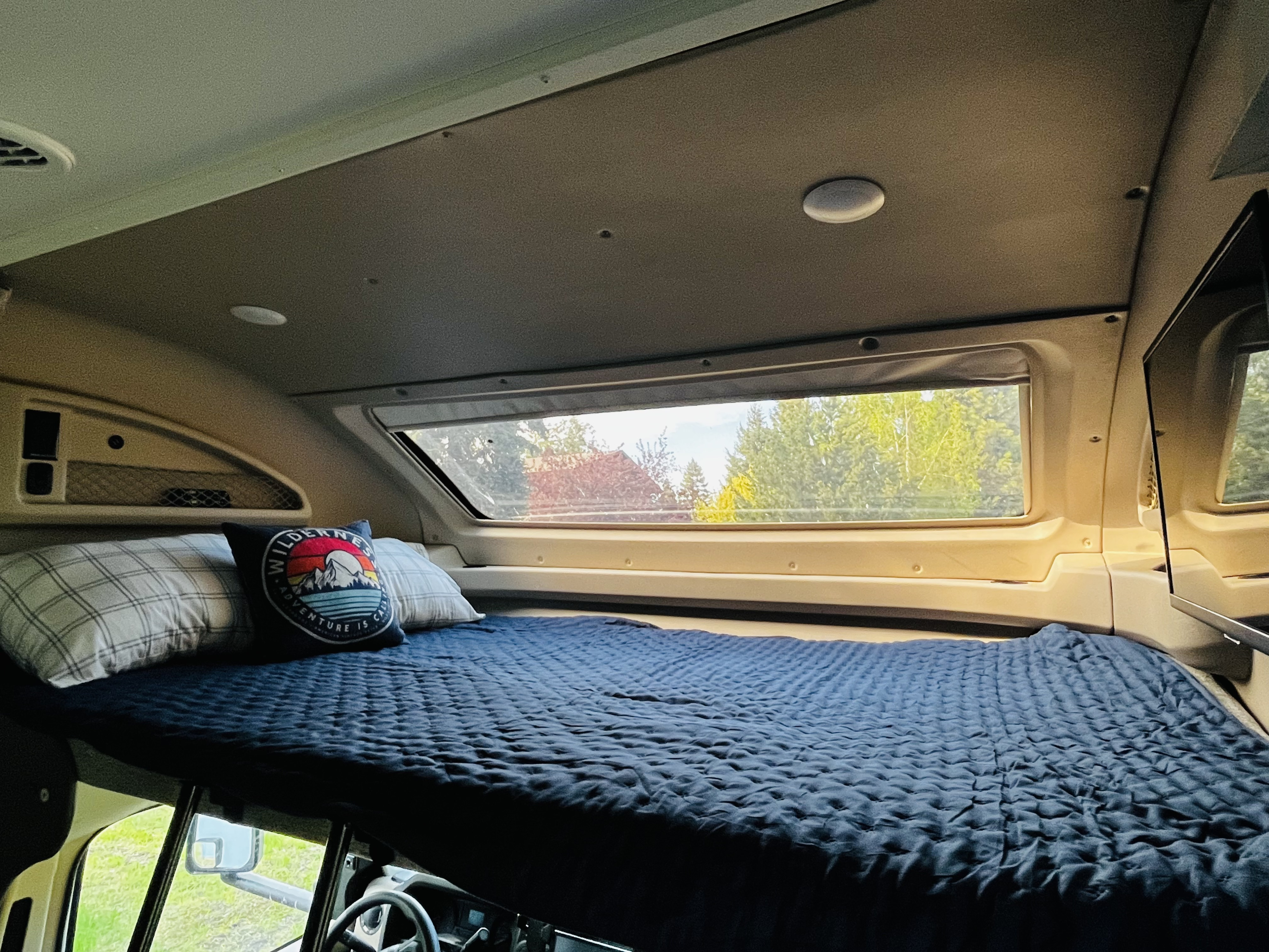Relax in your rv rental's cozy cab-over bed with a quilted blue blanket, pillows, and a scenic window view of trees.. Entegra Coach Odyssey 2021