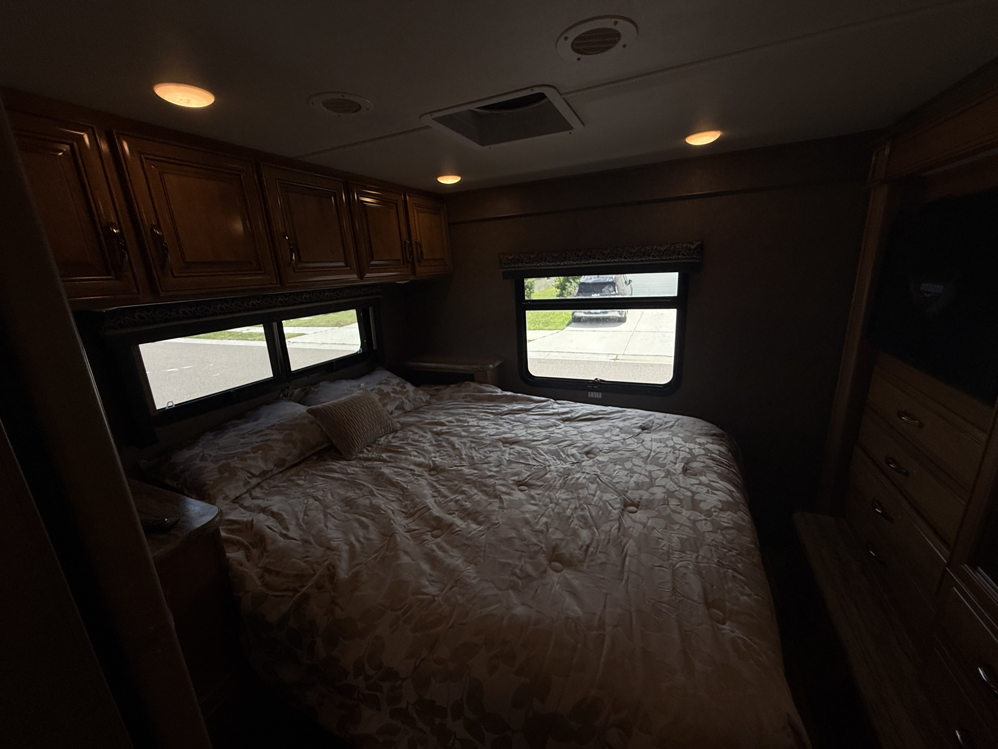 Dark, yet inviting RV rental bedroom. Features a large bed, wooden cabinets, and bright windows with exterior views, lit by warm ceiling lights.. Thor Quantum 2017