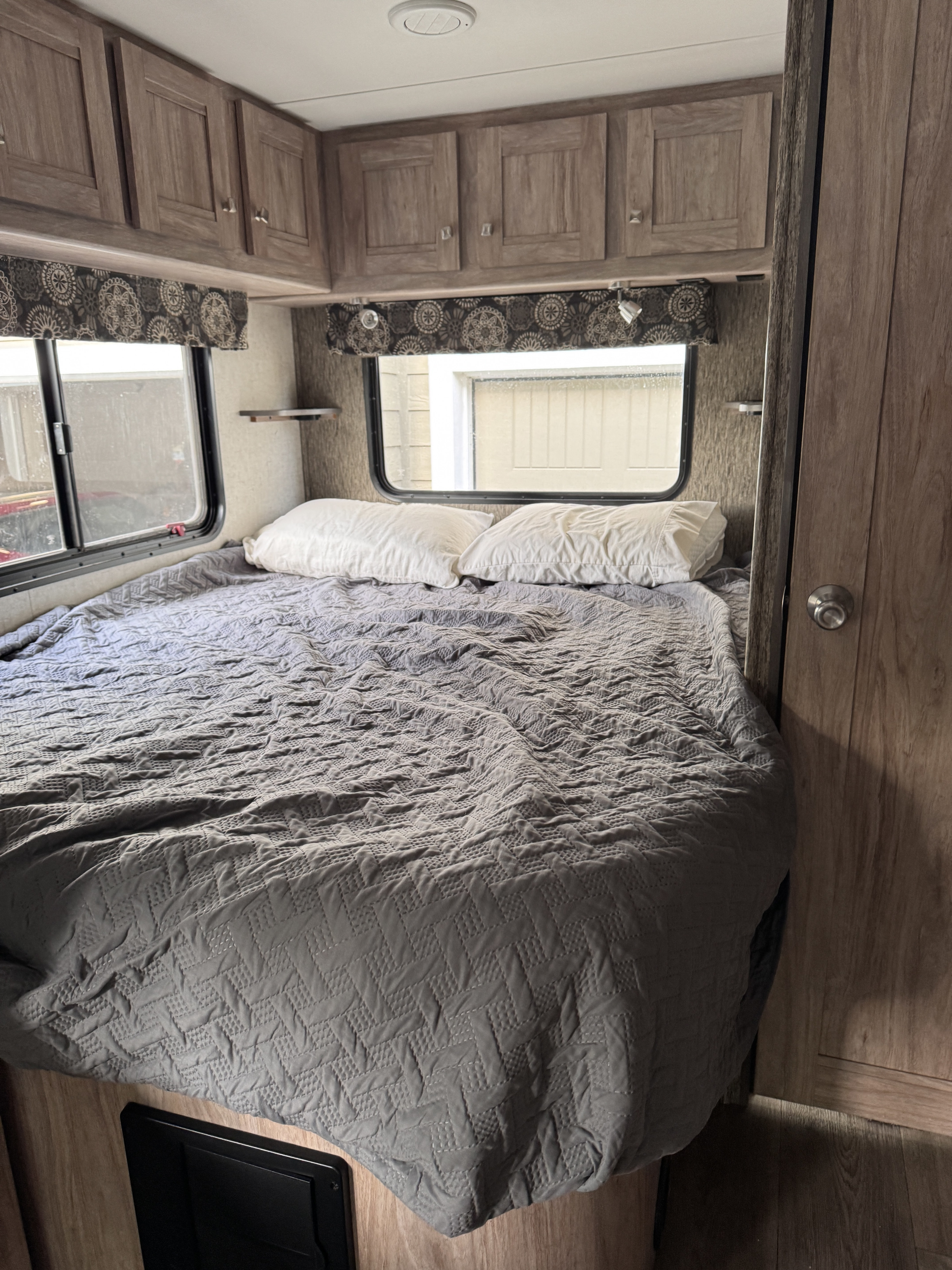 Cozy RV rental bedroom featuring a grey quilted bed, white pillows, overhead cabinets, and a window for natural light.. Forest River Sunseeker 2018