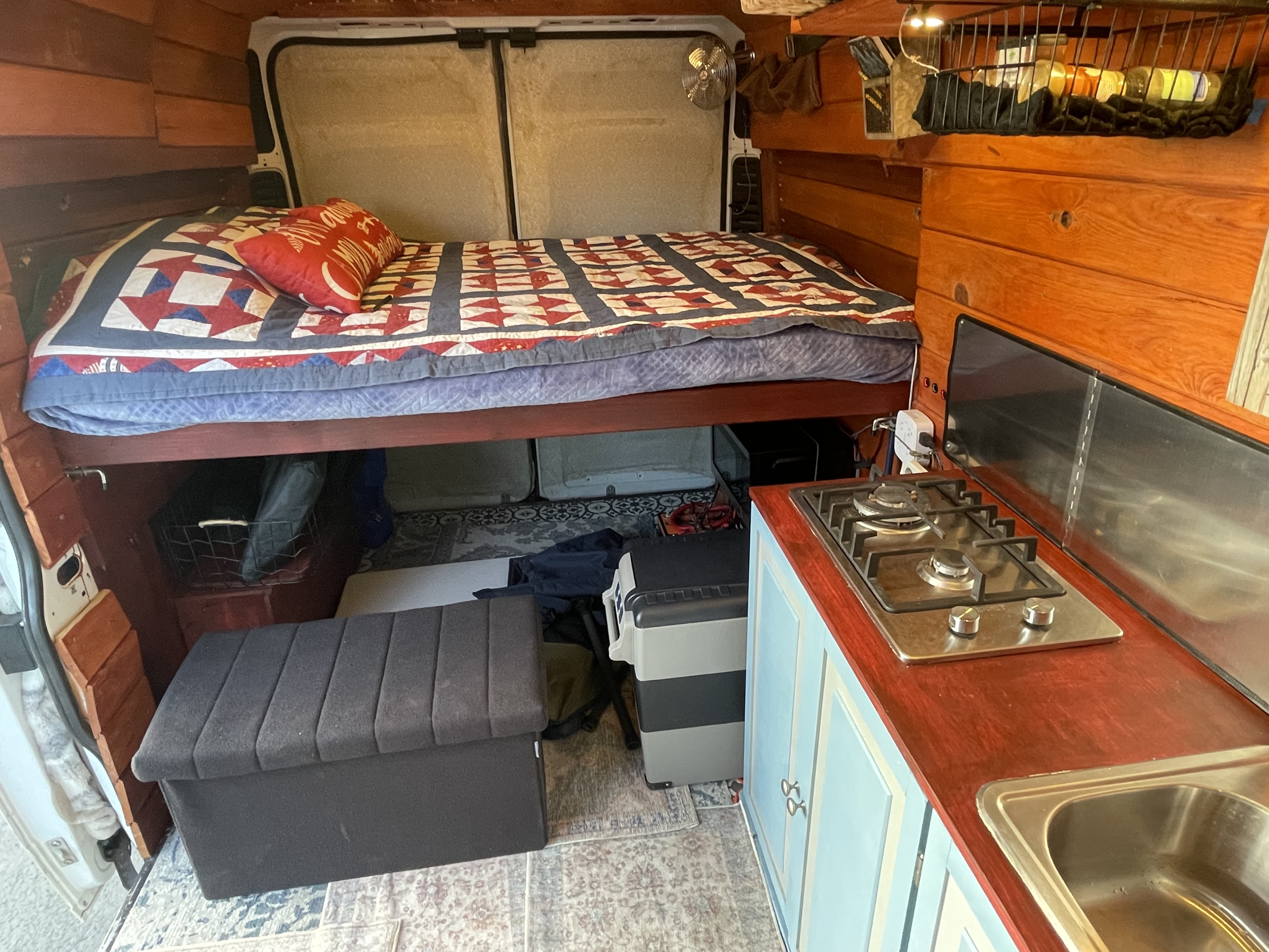 This RV rental interior features a cozy bed, compact kitchen with a stovetop/sink, wooden walls, & storage, ideal for road trips.. Dodge Other 2021