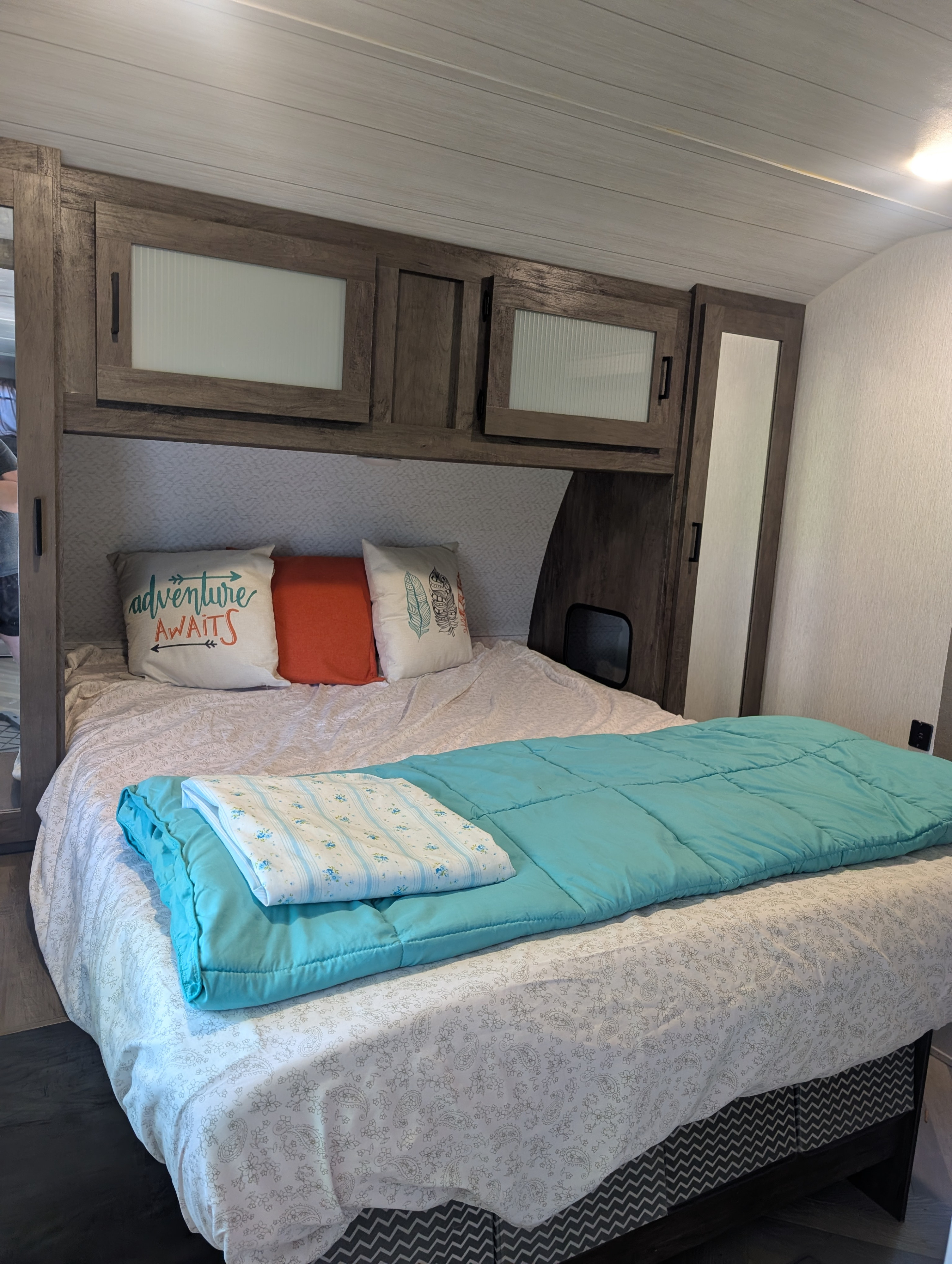 Experience comfort in this RV rental bedroom: cozy bed with turquoise comforter, adventure pillows, and ample overhead storage.. Forest River Wildwood 2022