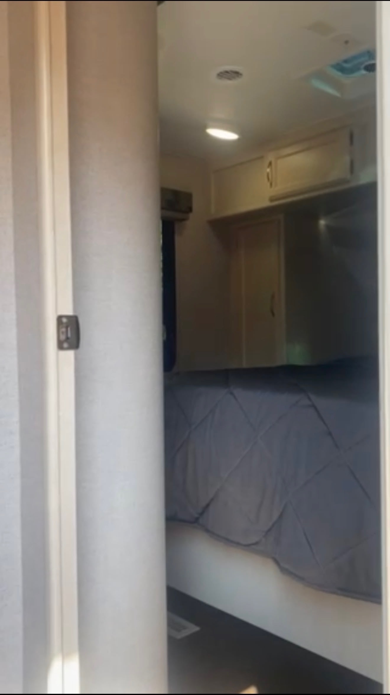 An RV rental bedroom interior, seen through an open door, features a dark quilted bed, overhead cabinets, and ceiling lights.. Winnebago 2500RL 2018