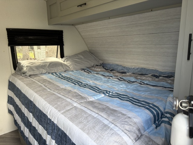 RV rental bedroom with blue, grey & white striped bed, pillows, window view, and overhead storage. Ready for travel.. Forest River Wildwood FSX 2022