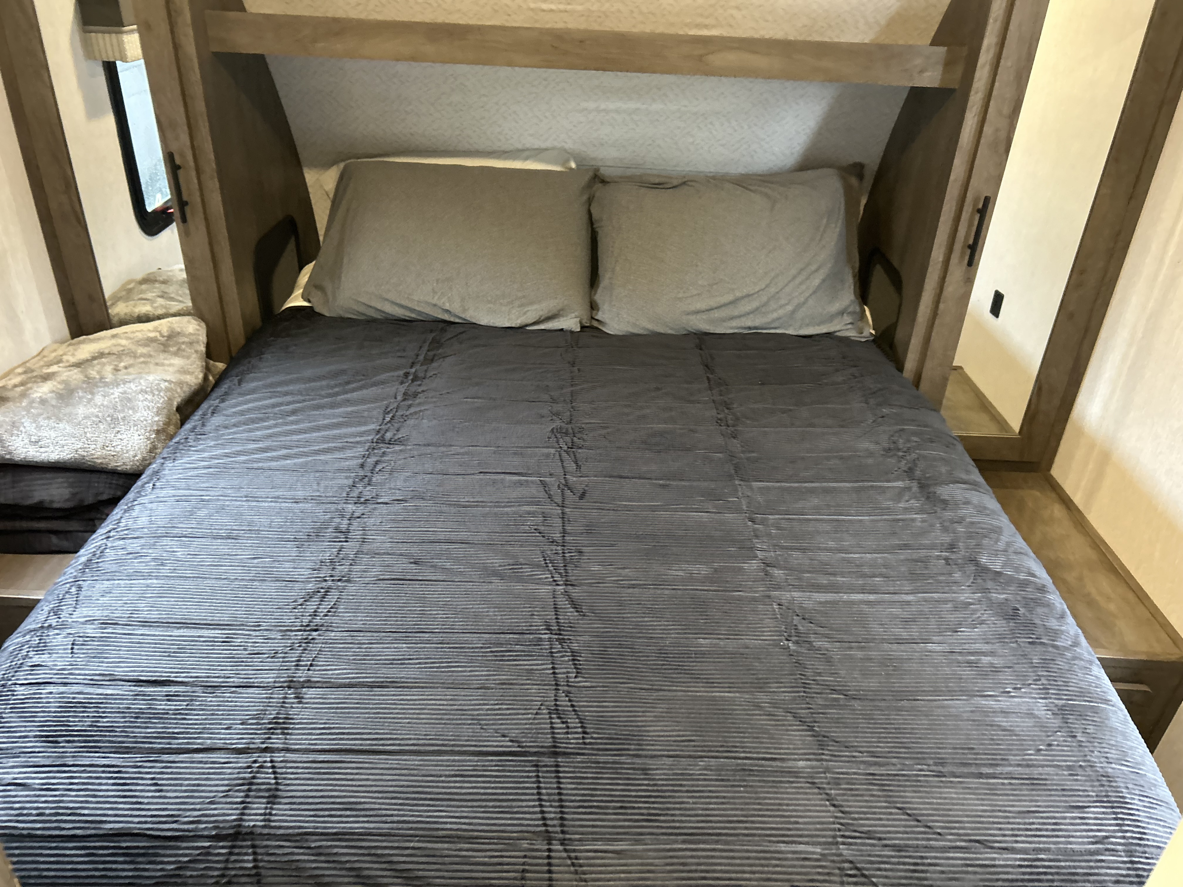 Cozy RV rental bedroom featuring a bed with gray comforter and pillows, nestled in a compact, wood-paneled alcove.. Forest River Wildwood X-Lite 2022