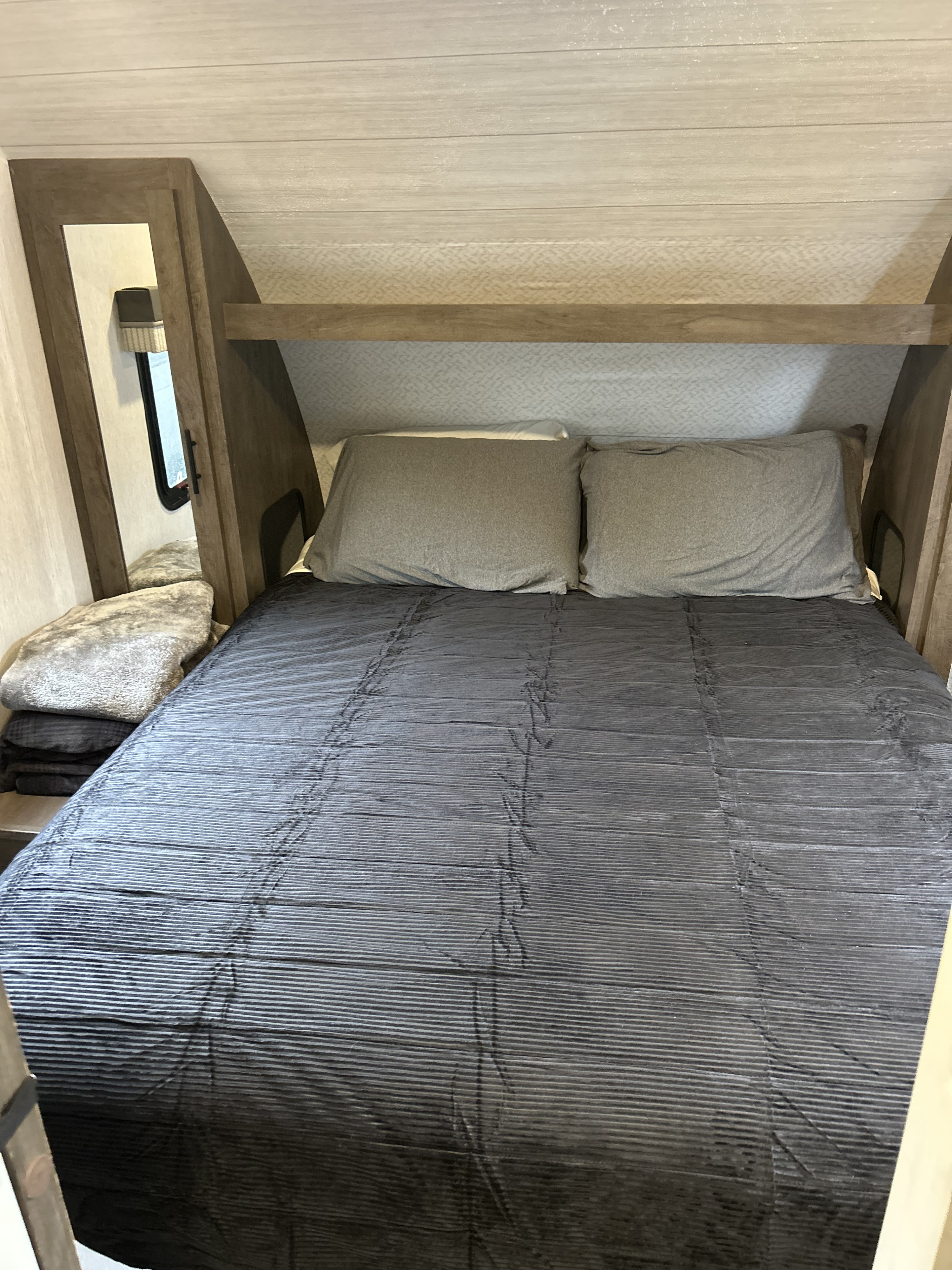 Cozy RV rental bedroom featuring a bed with a striped comforter, gray pillows, built-in storage, and a small window.. Forest River Wildwood X-Lite 2022