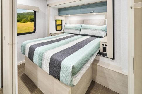 Inside a comfortable RV rental bedroom, featuring a striped bed, overhead cabinets, and a window with a green view.. Winnebago Sunflyer 26FE 2026