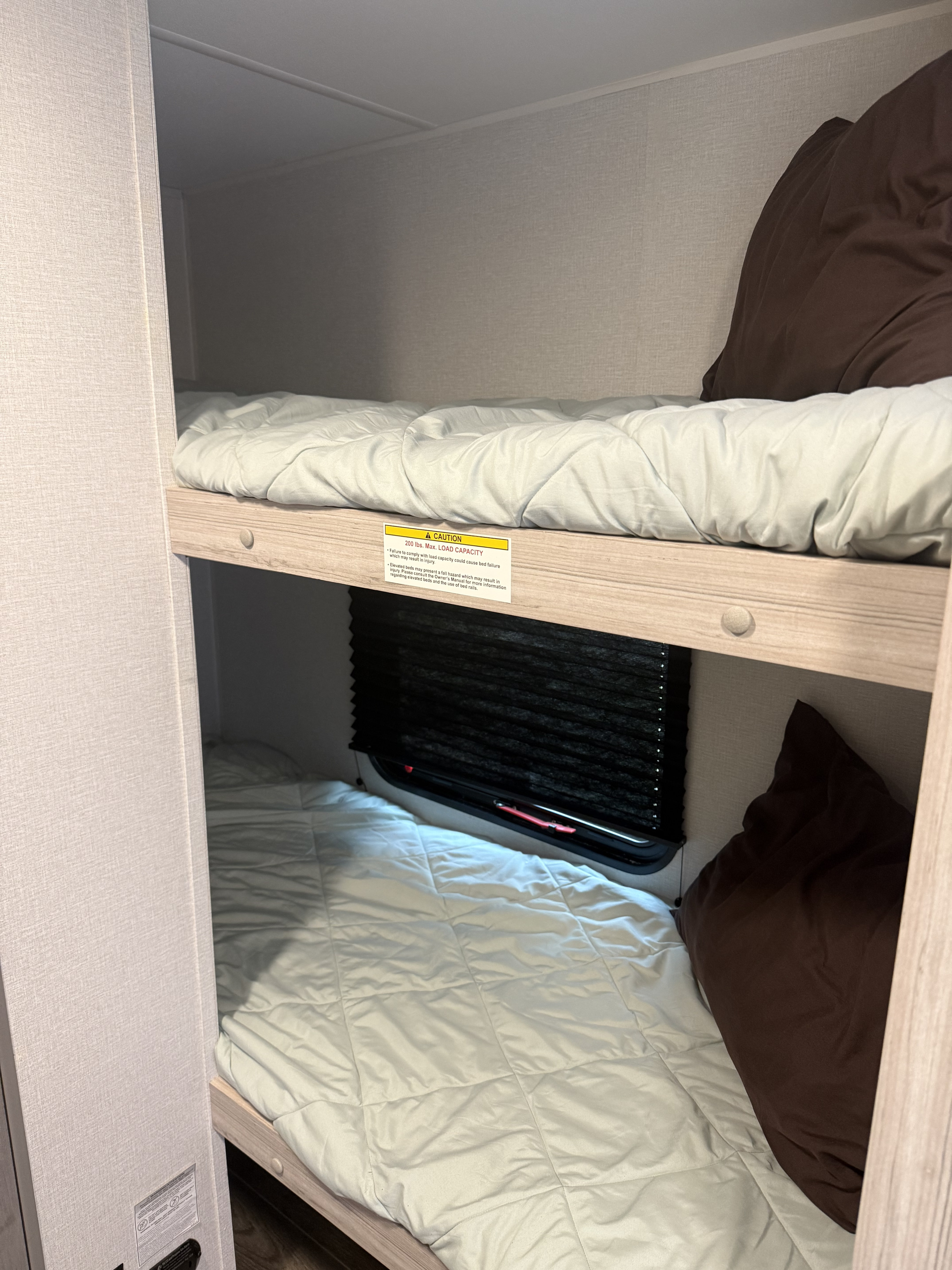 Cozy bunk beds in an rv rental, featuring light bedding, brown pillows, and a window with a black blind. A 200 lb weight limit sticker is visible.. Heartland RVs Pioneer 2025