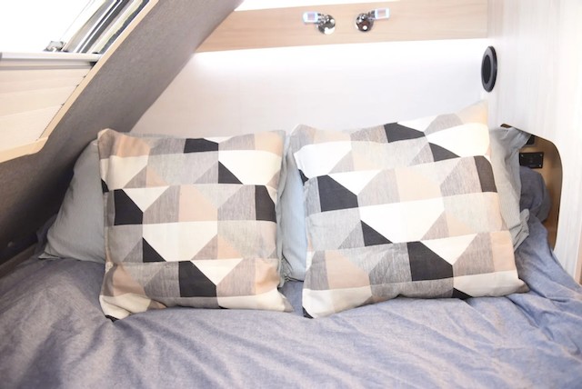 Cozy RV rental bed with stylish geometric pillows and light blue bedding, offering a comfortable sleeping area for travel.. nuCamp T@B 2024