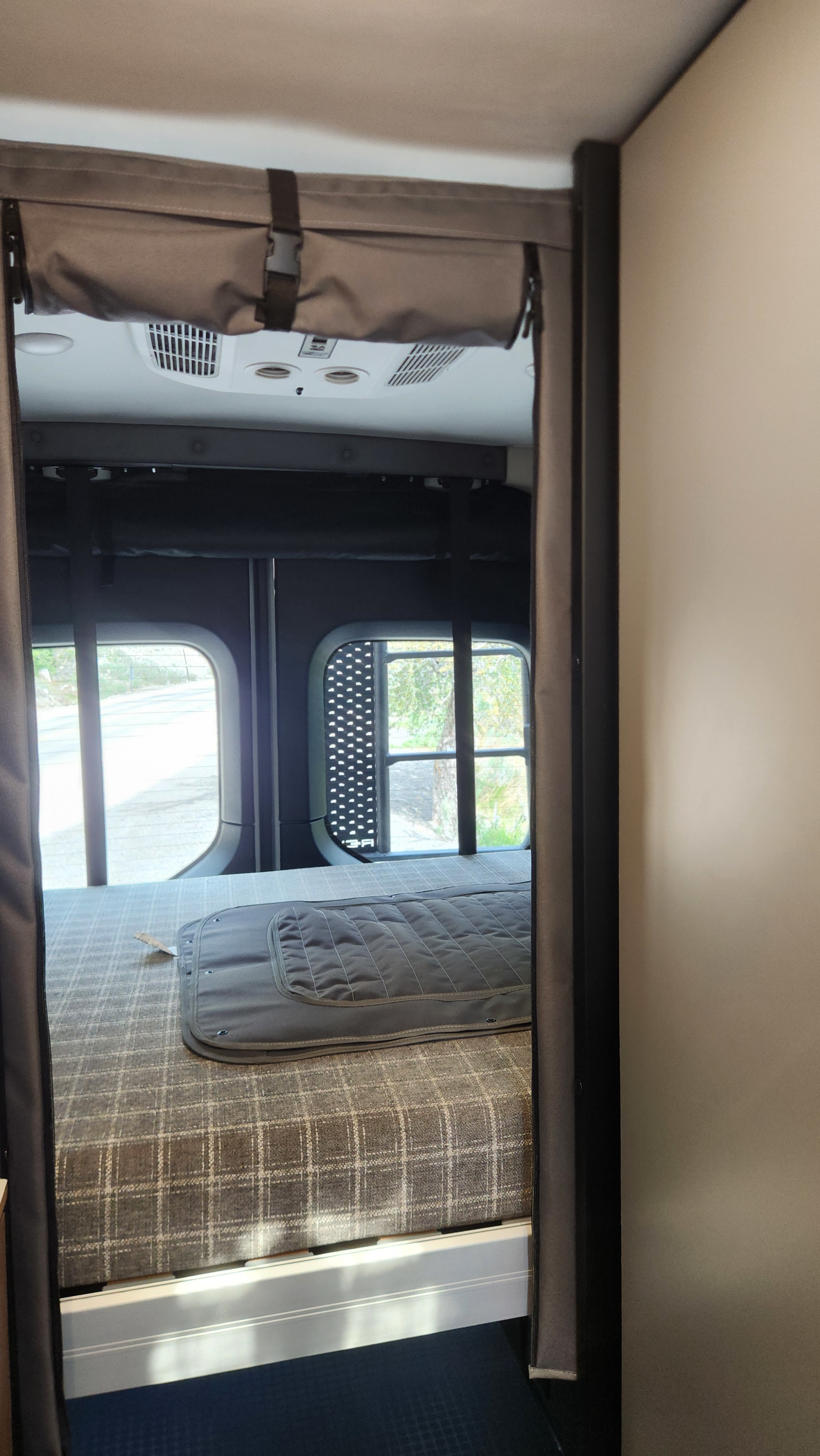 Experience comfort in this RV rental's bedroom. Features a plaid bed, large windows, and overhead AC in a compact camper van.. Winnebago Revel 2026
