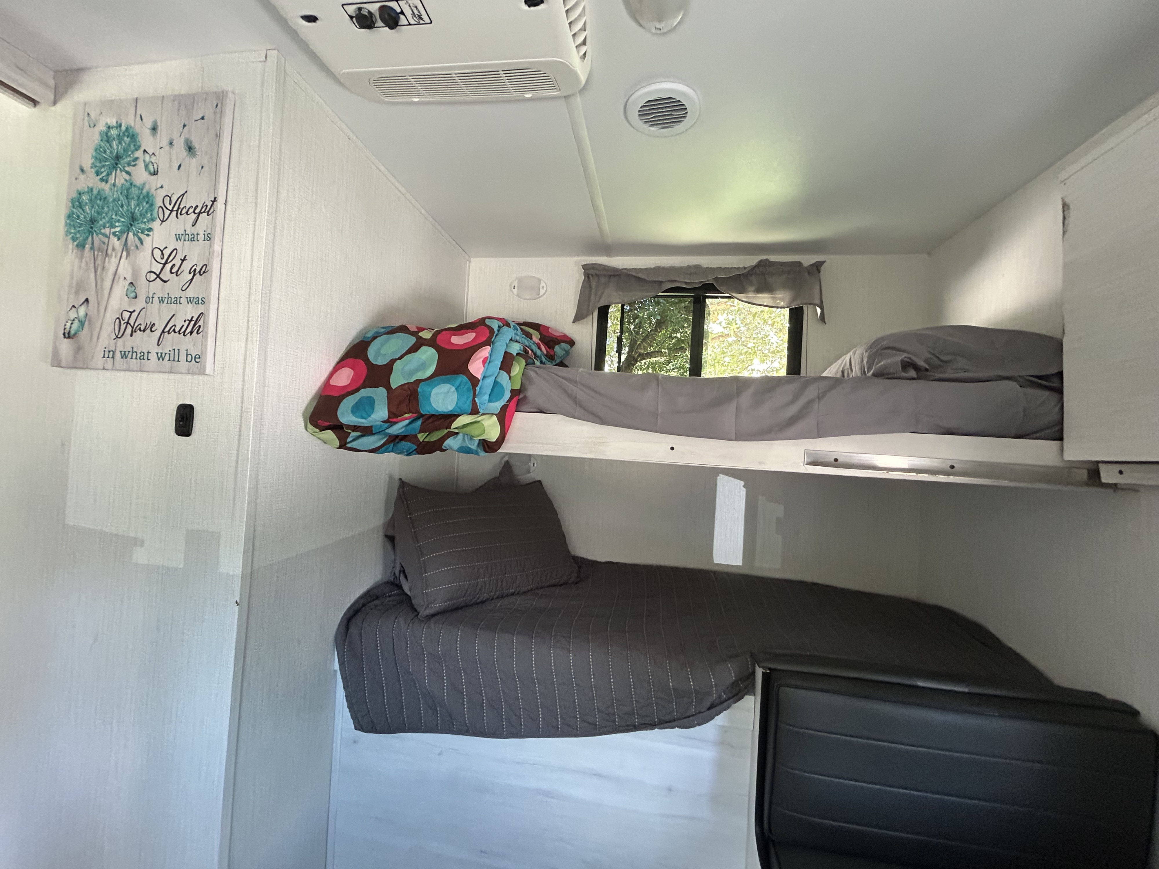 This RV rental interior features a cozy bunk bed setup with bedding, an AC unit, and a window overlooking trees.. Heartland RVs Trail Runner 2024