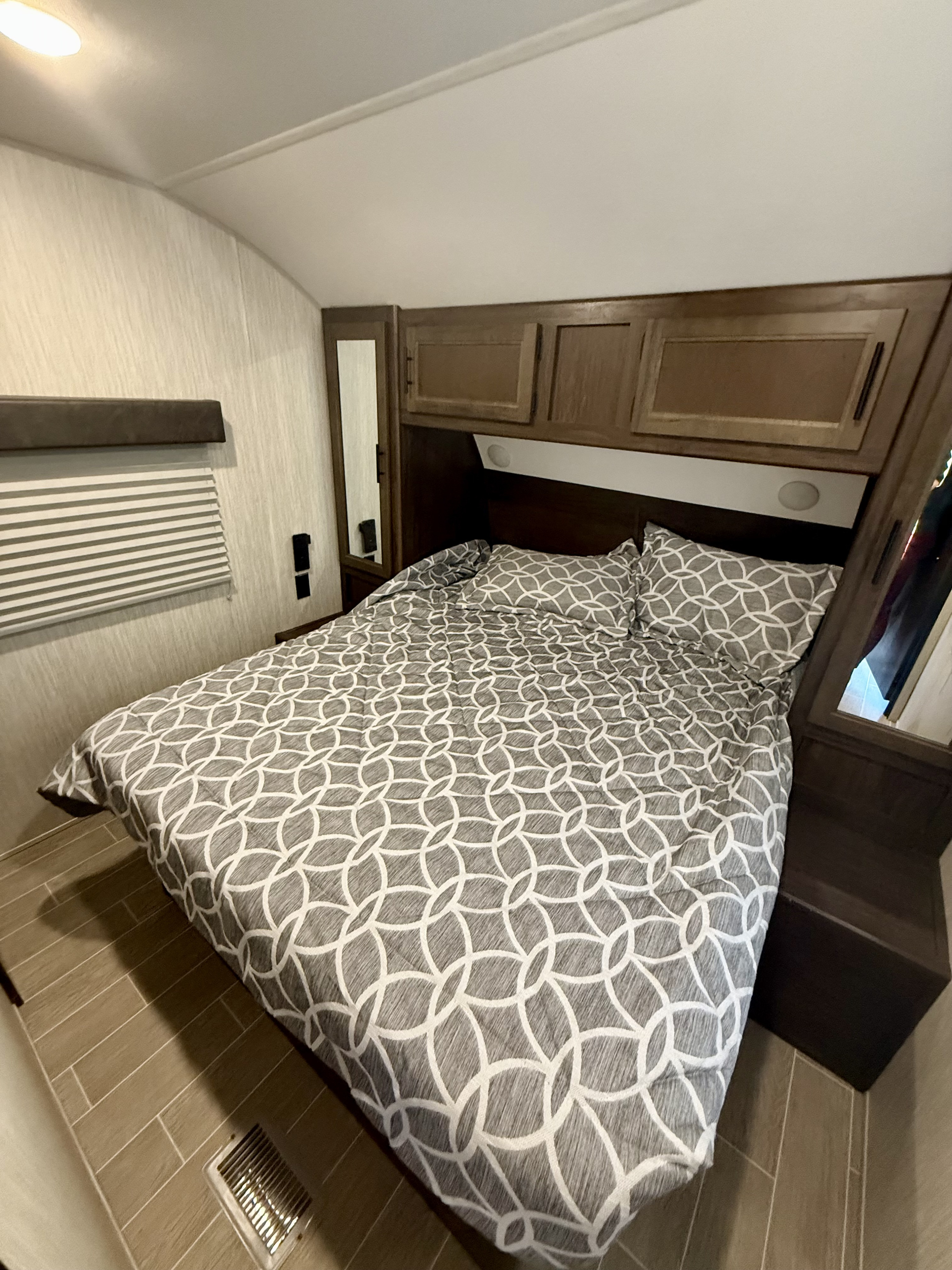 Cozy RV rental bedroom featuring a bed with gray patterned bedding, overhead cabinets, mirrors, window blinds, and wood-look tile flooring.. Forest River Cherokee 2021