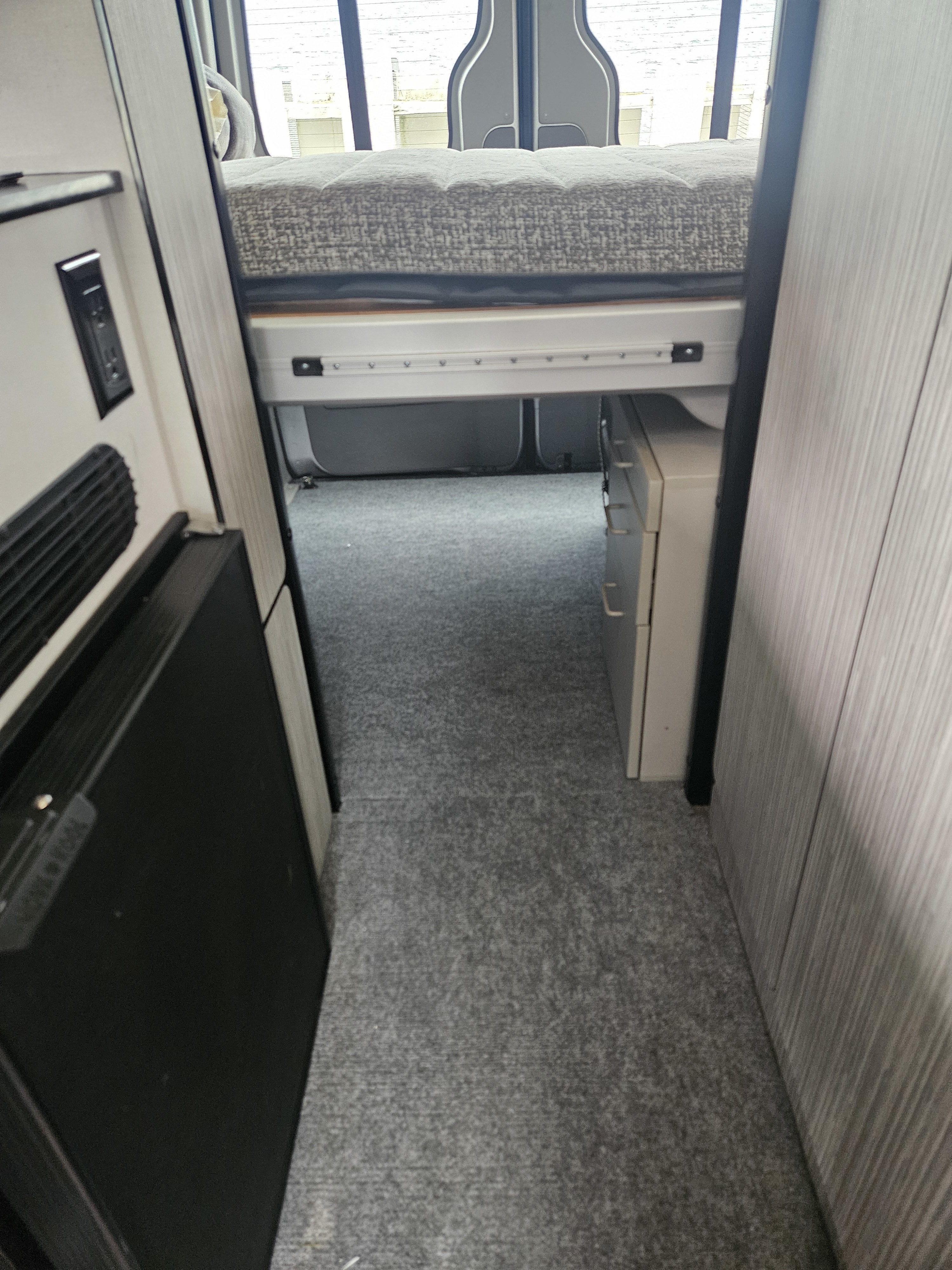 RV rental interior: elevated bed, under-bed storage, compact fridge on left, and a carpeted walkway.. Winnebago Revel 2018