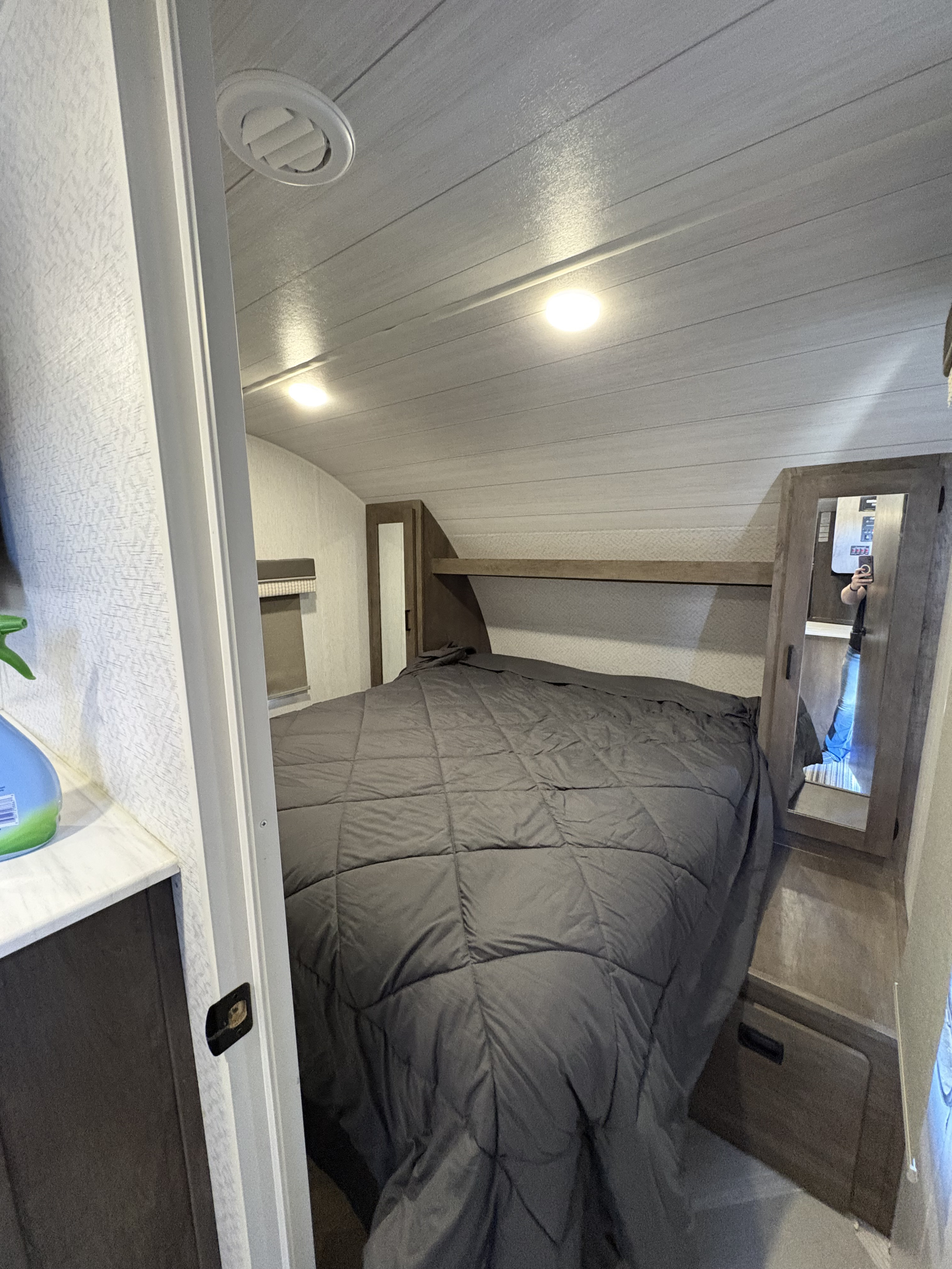RV rental bedroom: A dark grey bed with overhead storage. Mirrored cabinets and wood-look ceiling with lights create a compact, cozy space.. Forest River Wildwood X-Lite 2022