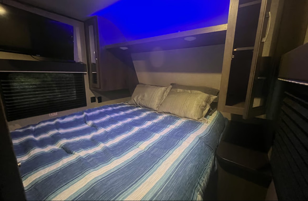 This RV rental's bedroom offers a cozy striped bed, mounted TV, window with blinds, and blue ambient lighting.. Jayco Jay Flight SLX 2021