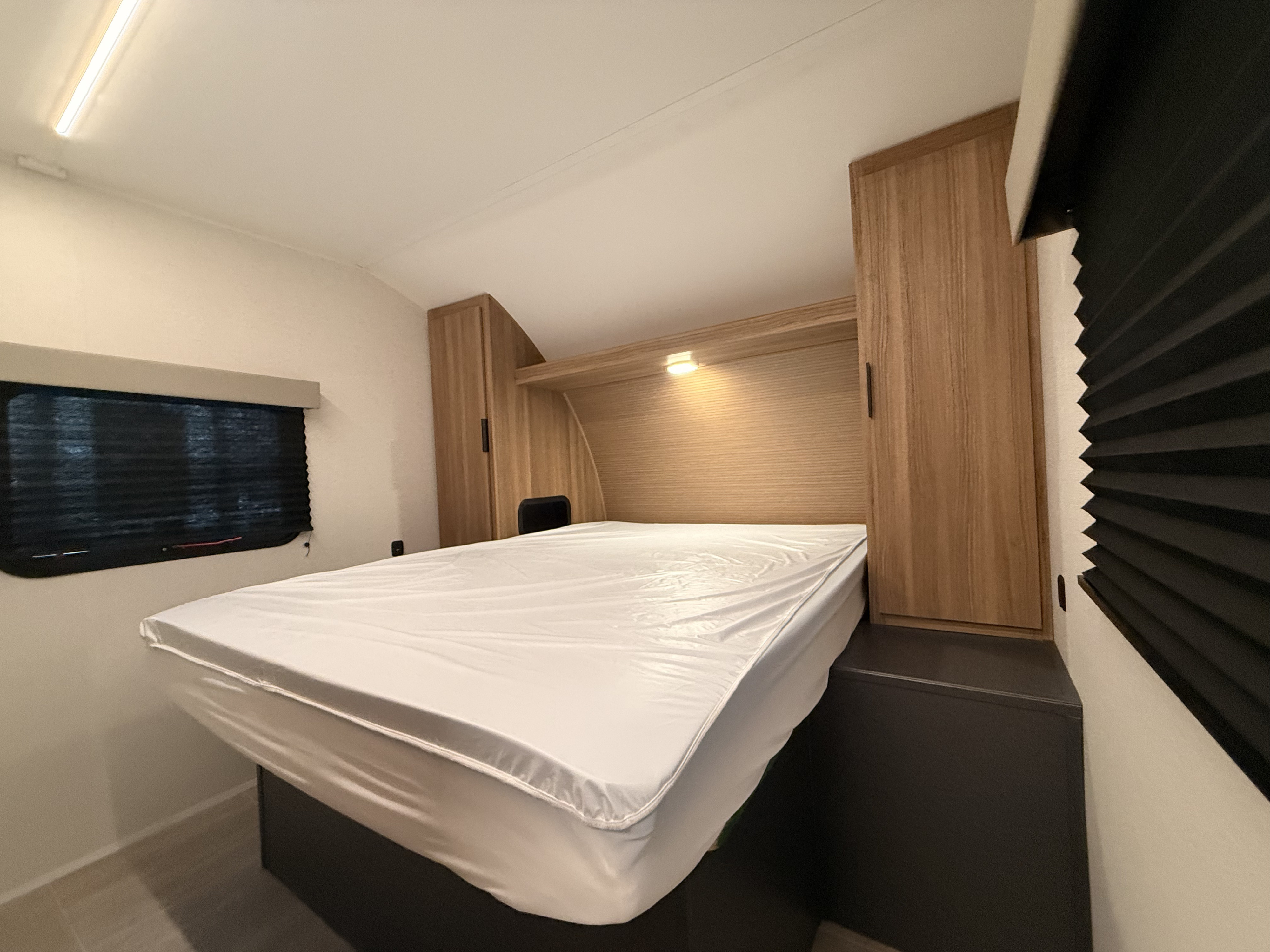 RV rental bedroom featuring a clean bed, wood storage, bright ceiling and reading lights, and dark window blinds.. Heartland RVs Trail Runner 2025