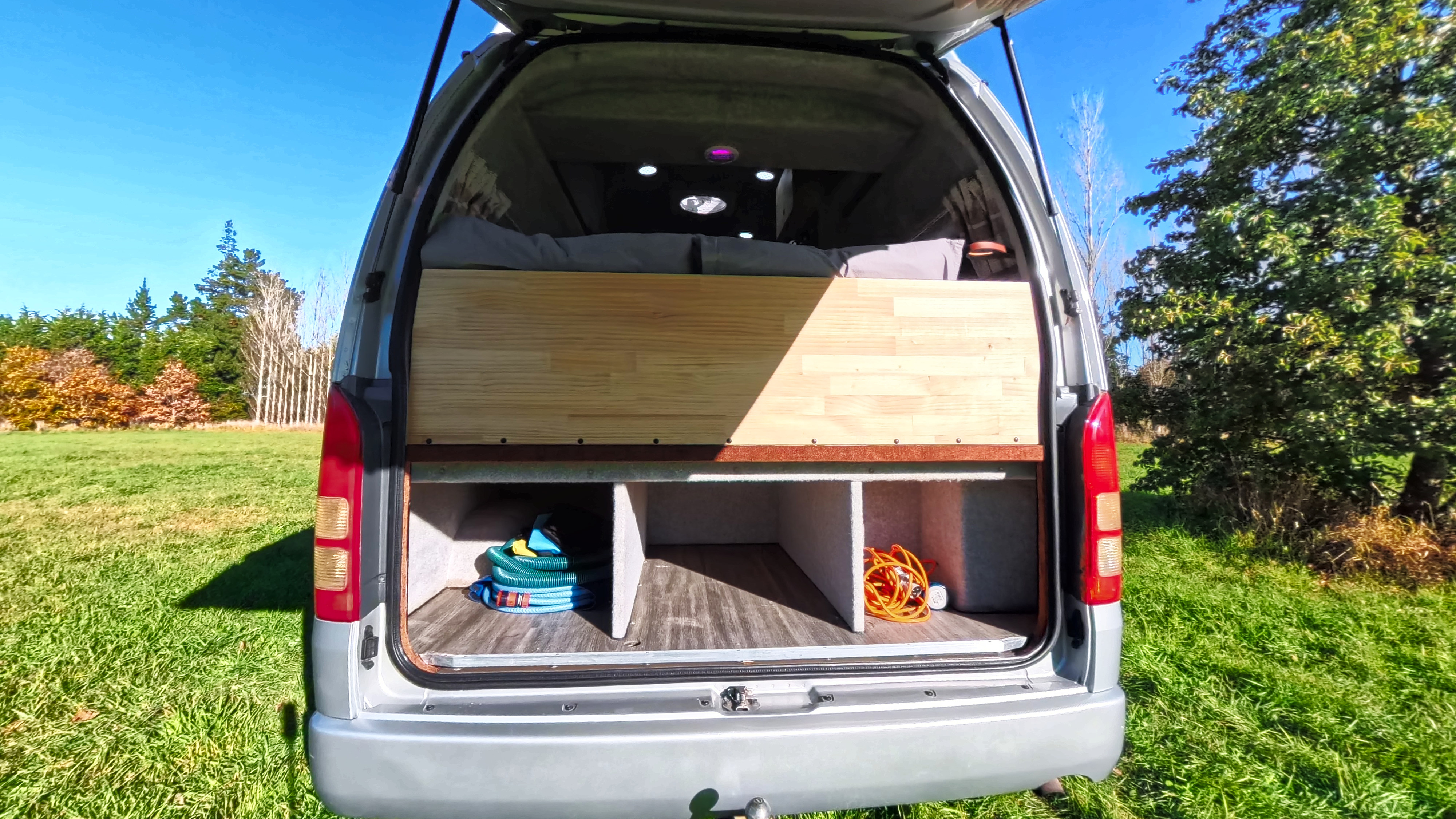 This RV rental's open tailgate reveals a comfy bed platform above storage. Parked in a sunny field with bright interior lights.. Other Other 2006