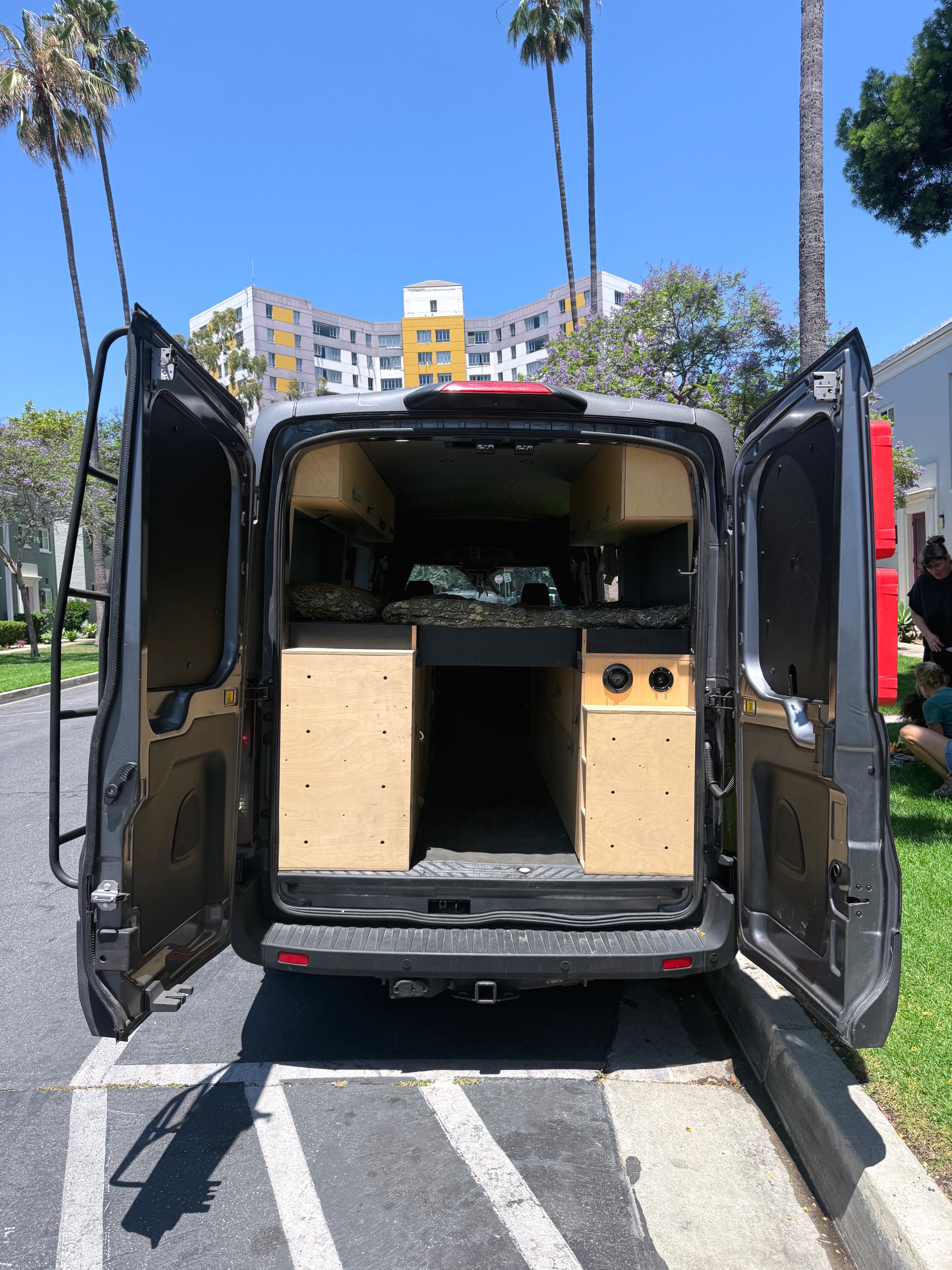 Custom RV rental campervan conversion with open rear doors revealing a bed, wood storage, and speakers. Parked outdoors on a sunny day.. Ford Transit 2021