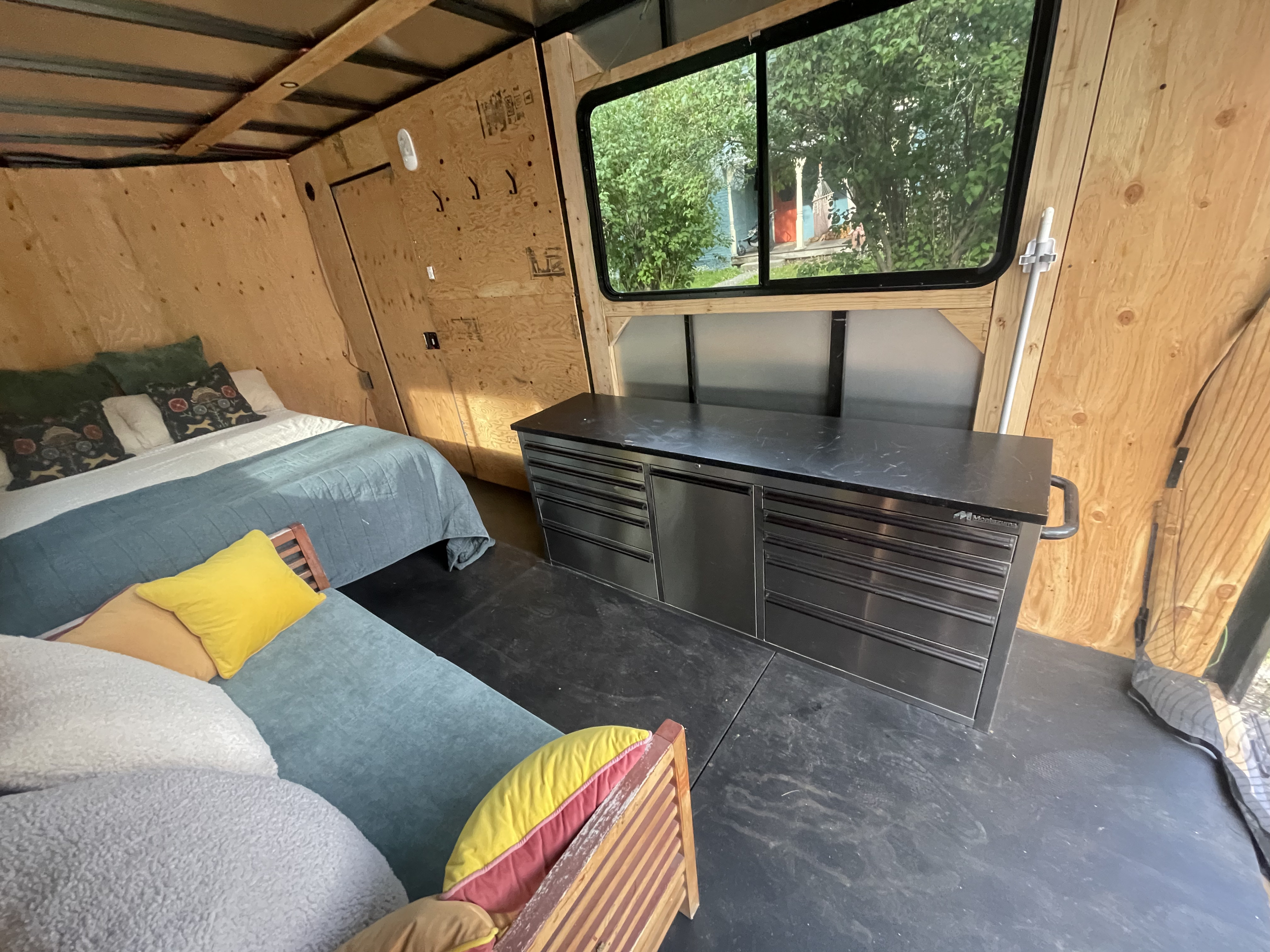 Rustic RV rental interior featuring a bed, blue couch, metal storage, and window with greenery. Plywood walls surround the space.. Royal Cargo Enclosed Trailer 2025