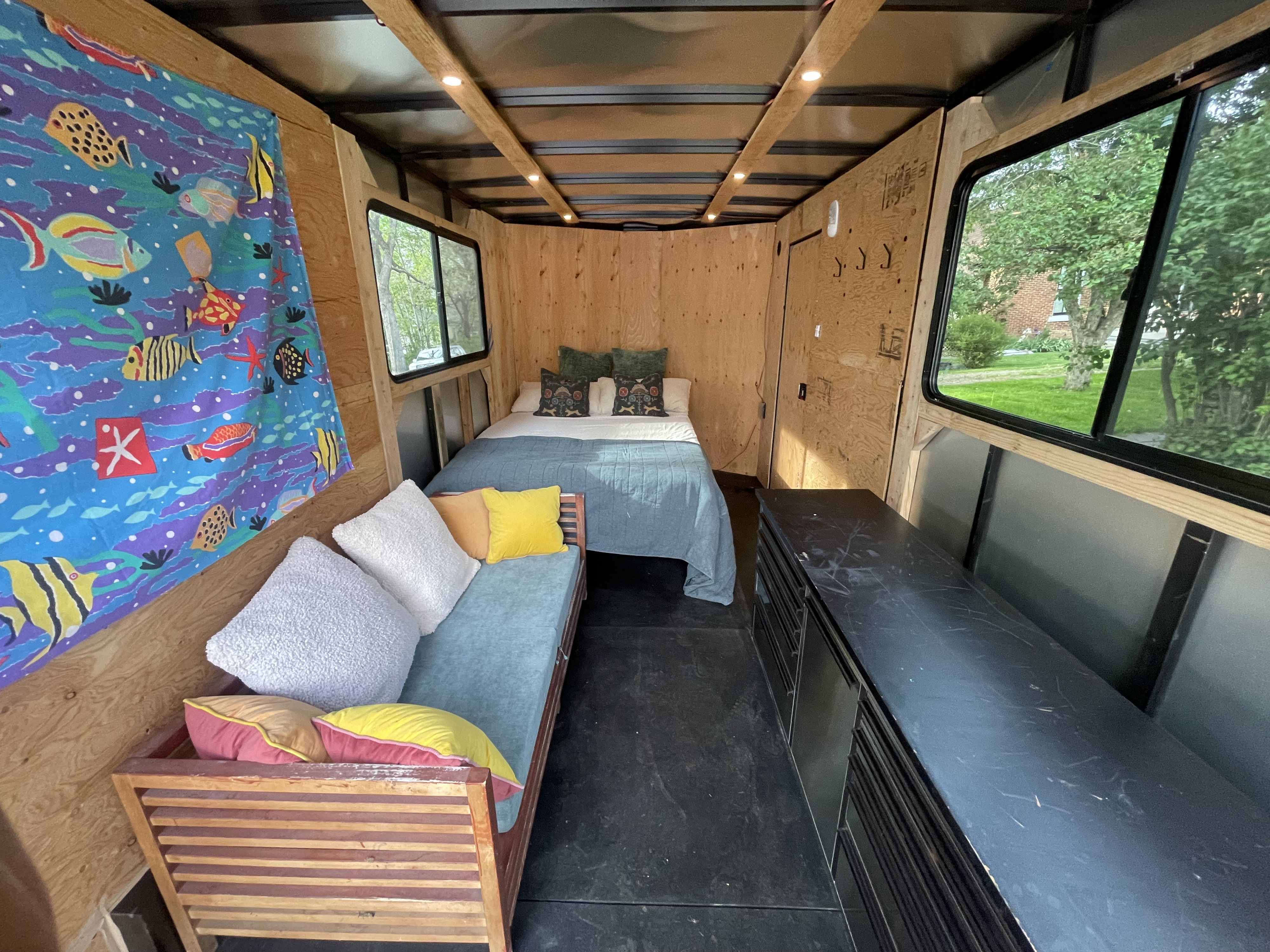 Cozy rv rental camper interior featuring a bed, vibrant couch, wood paneling, and outdoor views through windows.. Royal Cargo Enclosed Trailer 2025