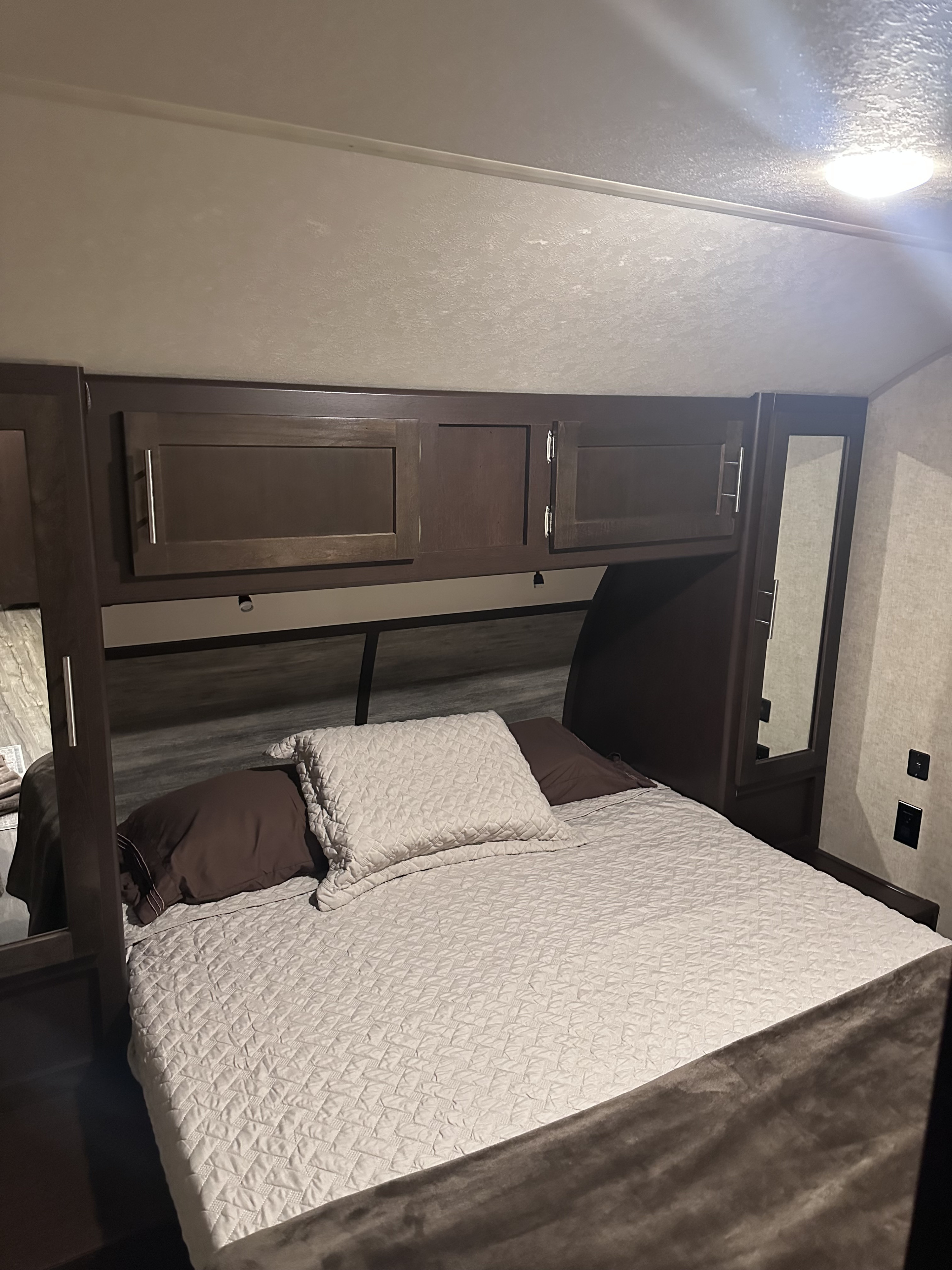 Cozy RV rental bedroom featuring a bed with a light-colored quilt, dark wood overhead cabinets, and a side mirror.. Forest River Cherokee 2017