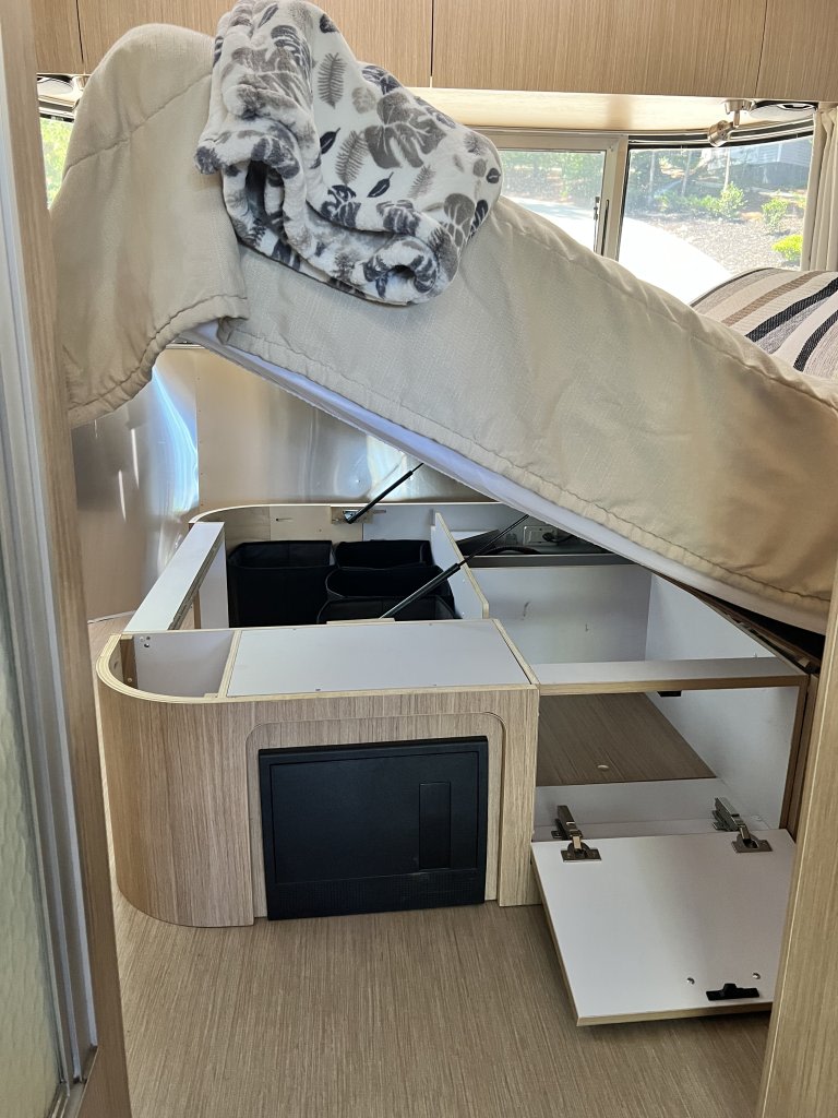 An rv rental bed is lifted, revealing under-bed storage with black bins, an electrical panel, and light wood cabinetry.. Airstream Flying Cloud 2018