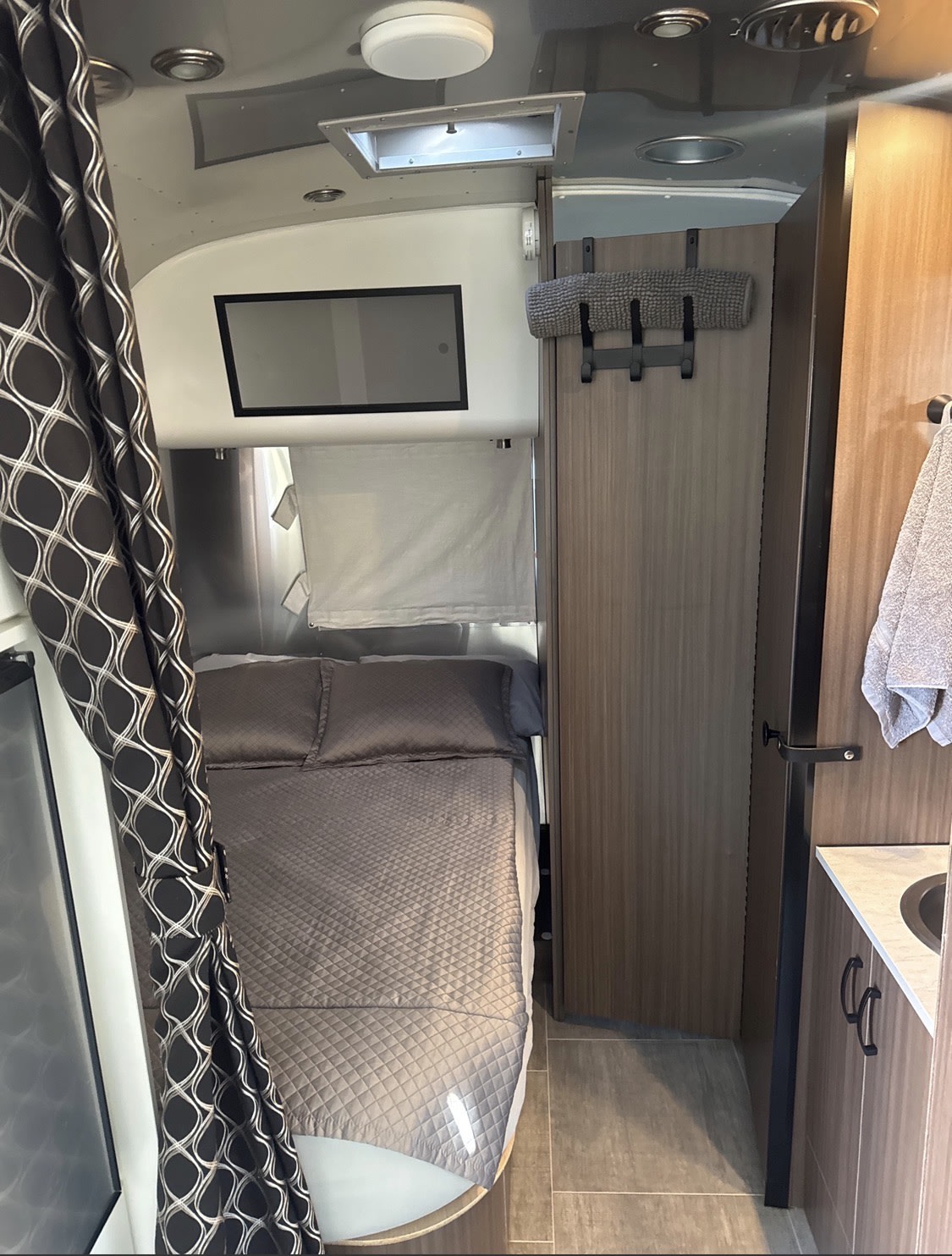 View inside an RV rental showing a small bedroom with gray bedding, an overhead vent, and a compact bathroom vanity.. Airstream Caravel 2020