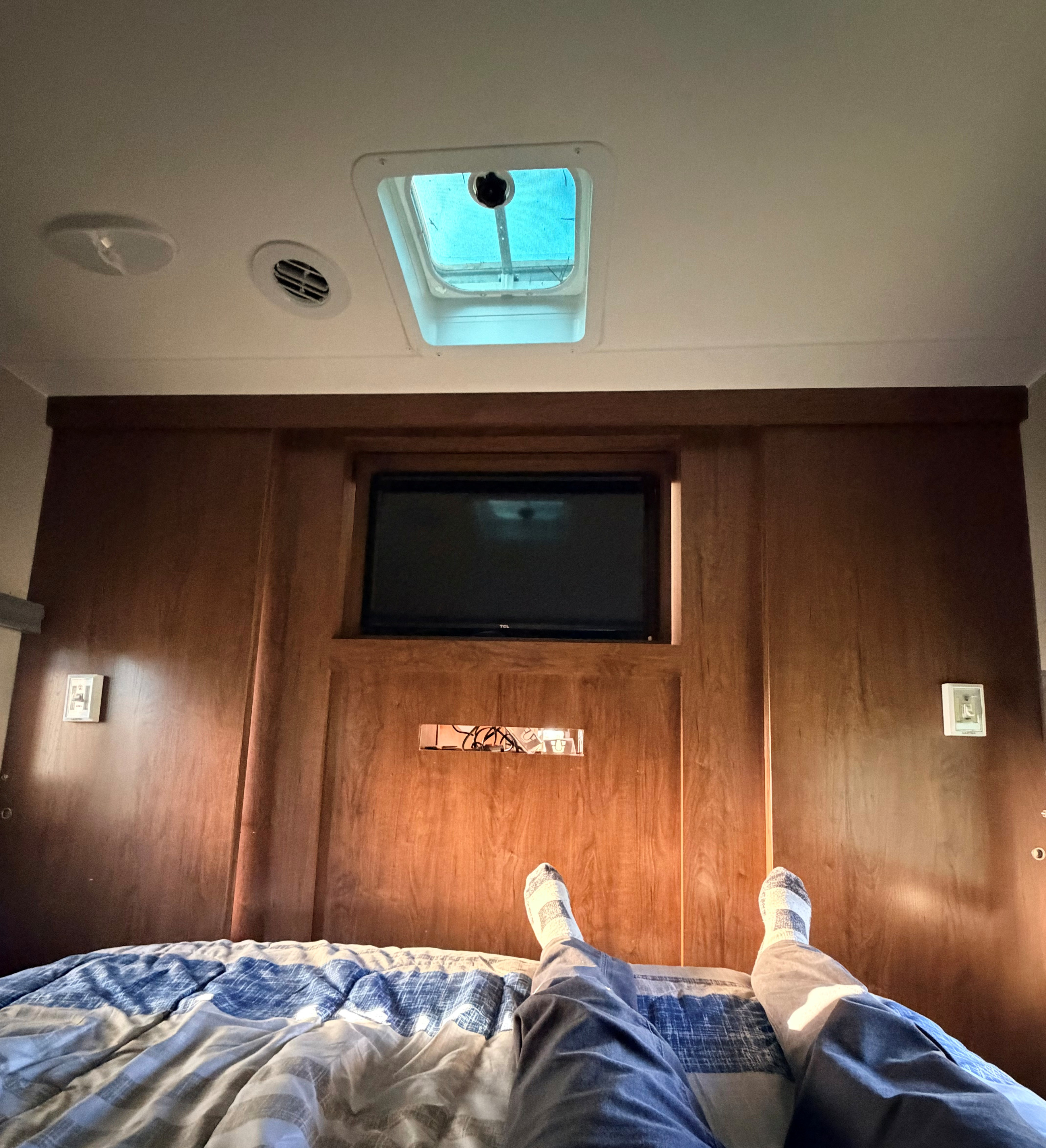 Relaxing in an RV rental: Legs on a blue bedspread, looking up at a TV embedded in a wooden wall, with a ceiling vent & skylight.. Coachmen Freedom Express 312BHDS Travel Trailer 2017