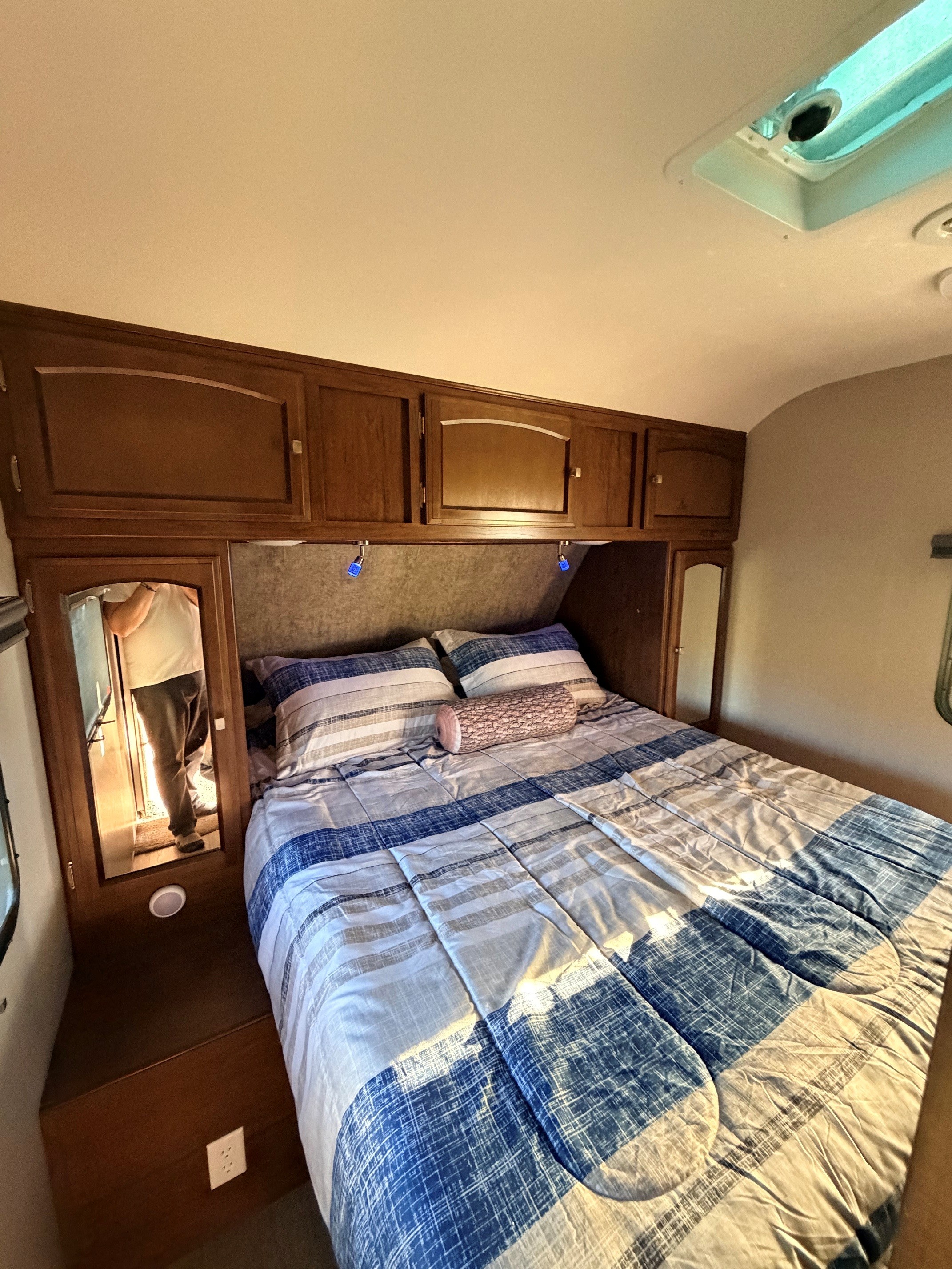 Cozy RV rental bedroom with striped bedding, log pillow, wood cabinets, and a mirror showing a person.. Coachmen Freedom Express 312BHDS Travel Trailer 2017