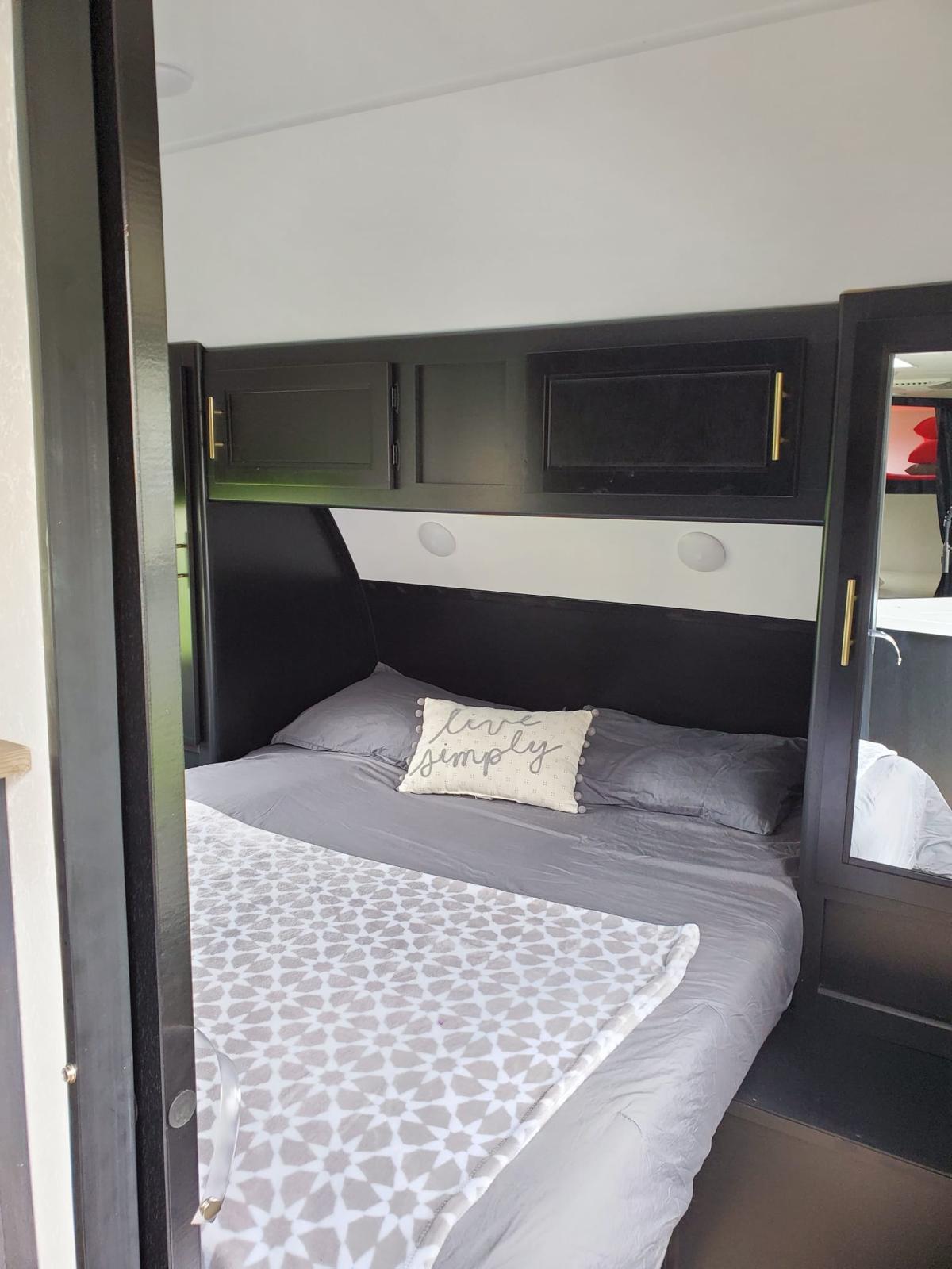 Modern RV rental bedroom: black overhead cabinets, gray bedding, a "live simply" pillow. Cozy and inviting space.. Forest River Cherokee Grey Wolf 2021