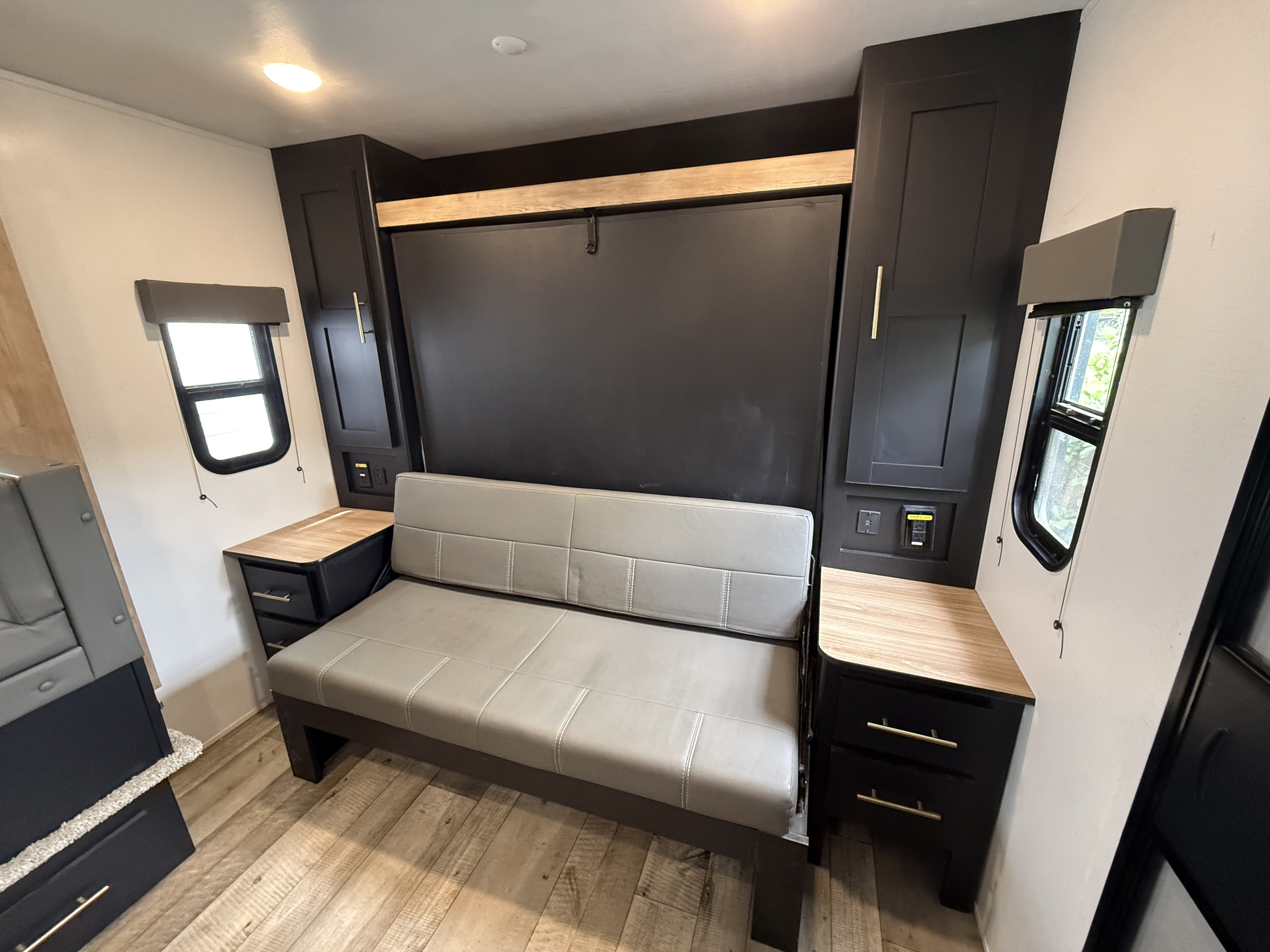 This RV rental interior showcases a cozy gray sofa, murphy bed, modern dark cabinetry, and wood-look flooring for compact living.. Keystone RV Springdale 2022